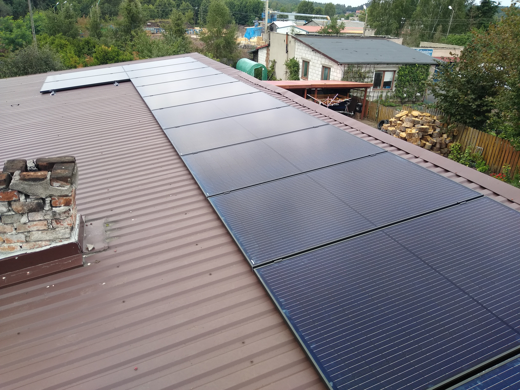 Rząd paneli słonecznych na brązowym, metalowym dachu domu jednorodzinnego, widok z góry, w tle zabudowania i drzewa.