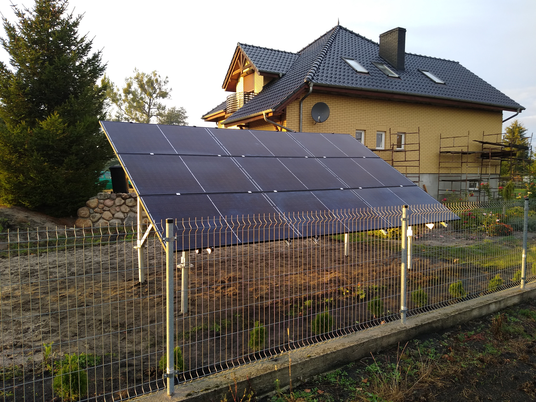 Instalacja fotowoltaiczna na gruncie w Poznaniu, widoczna za metalowym ogrodzeniem, na tle domu z żółtej cegły i rusztowaniami.