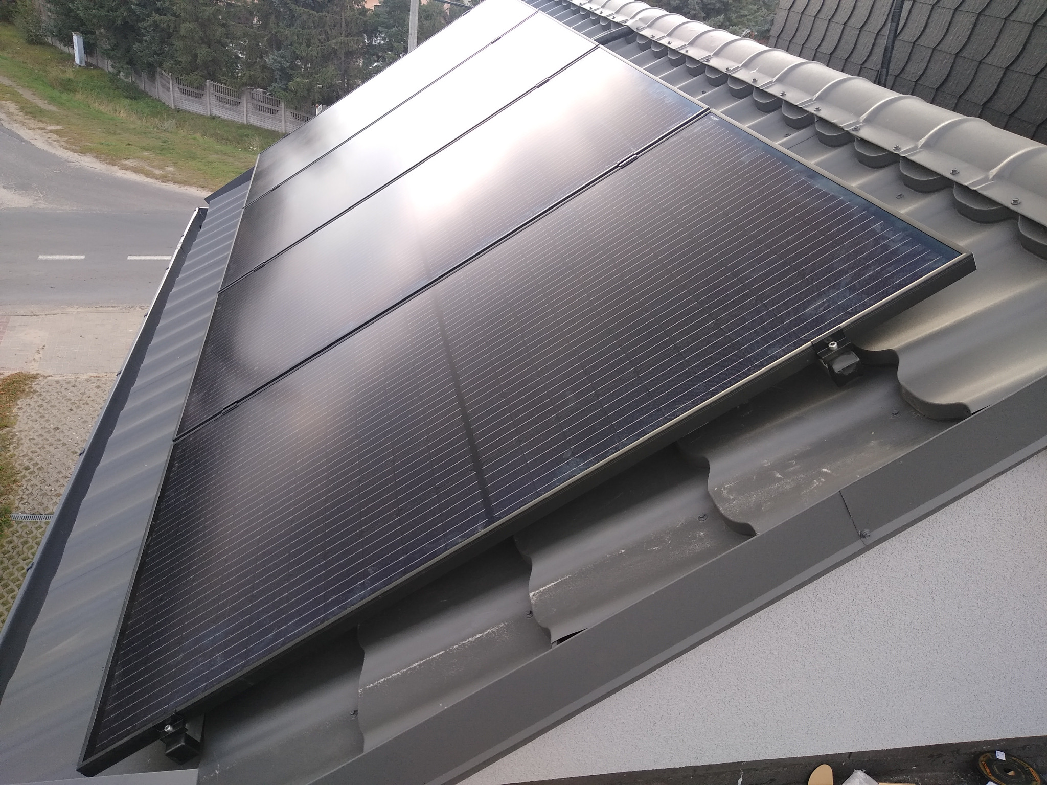 Cztery czarne panele słoneczne zamontowane na szarym dachu z dachówki ceramicznej, widok z góry pod kątem, w tle ulica i drzewa.