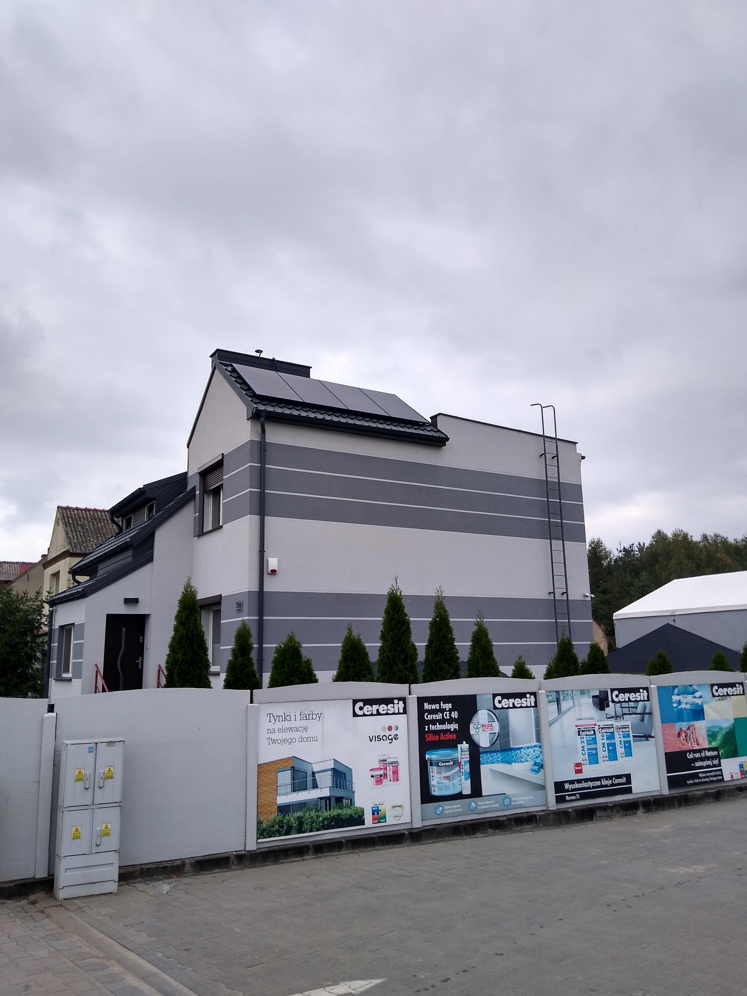 Dwukondygnacyjny dom z panelami słonecznymi na dachu, widoczny z poziomu ulicy, z reklamami materiałów budowlanych na pierwszym planie.