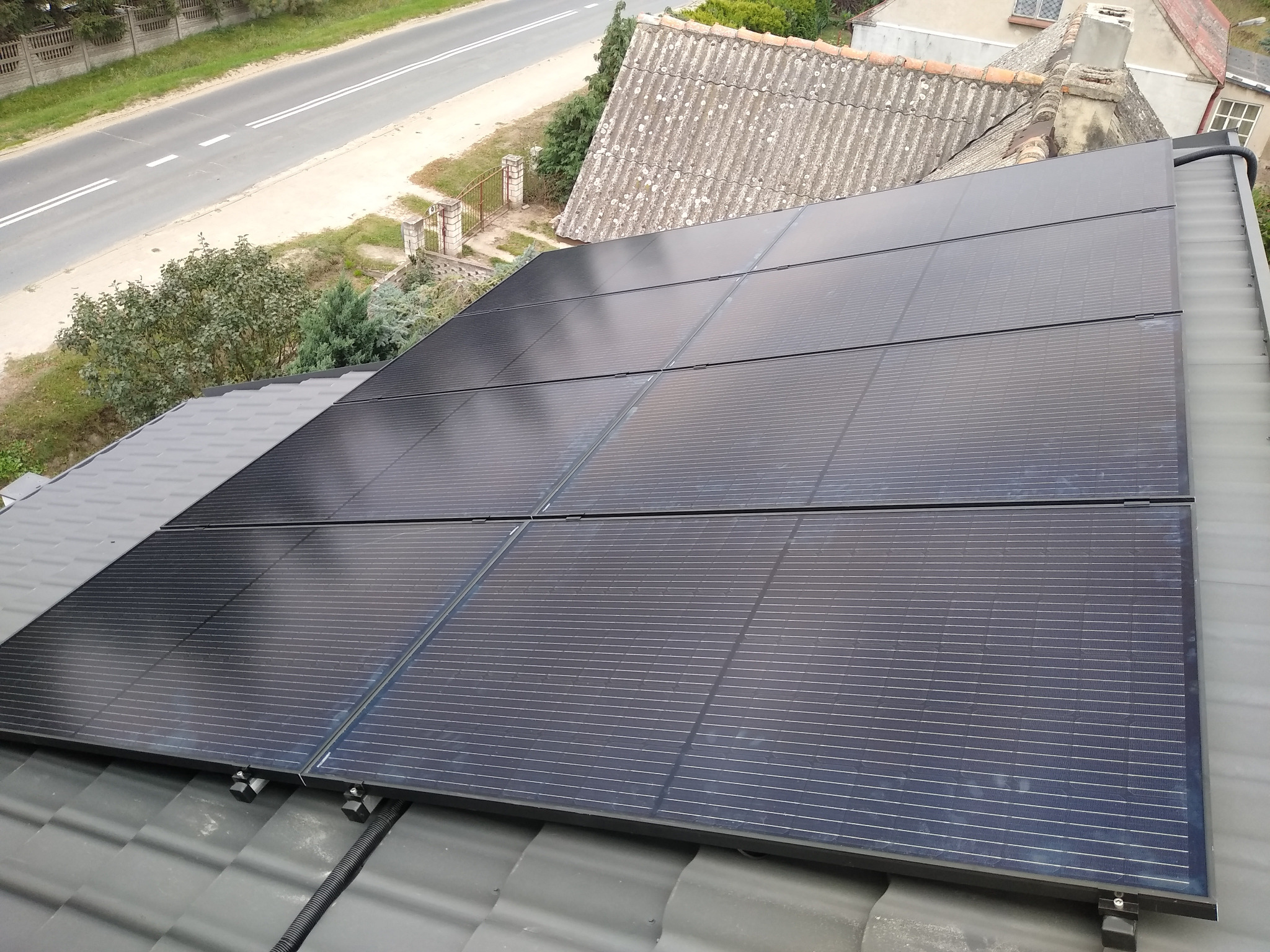 Czarny panel słoneczny na szarym dachu z widokiem na drogę i dom z azbestowym dachem w tle.