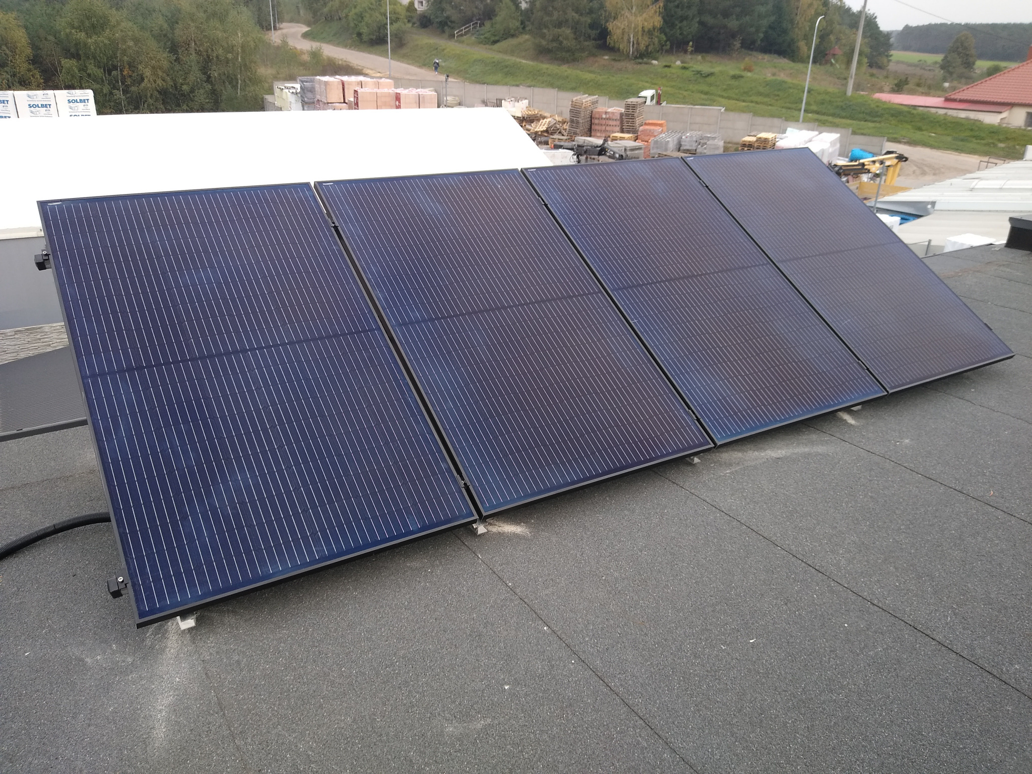 Cztery panele słoneczne na płaskim dachu pokrytym papą, widoczne elementy montażowe i przewody, w tle budynki i drzewa.