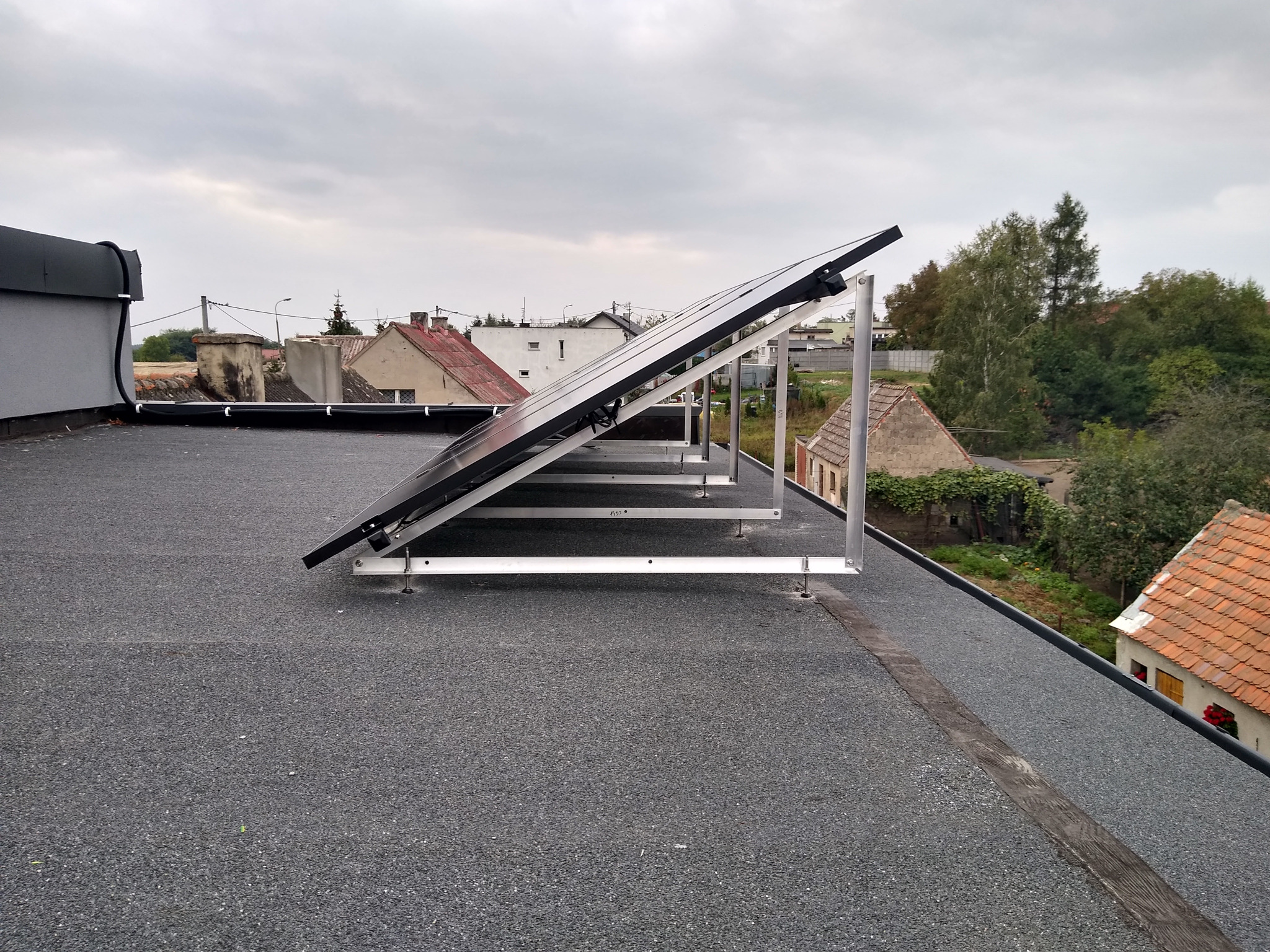 Panele słoneczne zamontowane na płaskim dachu pokrytym papą, widok z boku, w tle zabudowa jednorodzinna i pochmurne niebo.