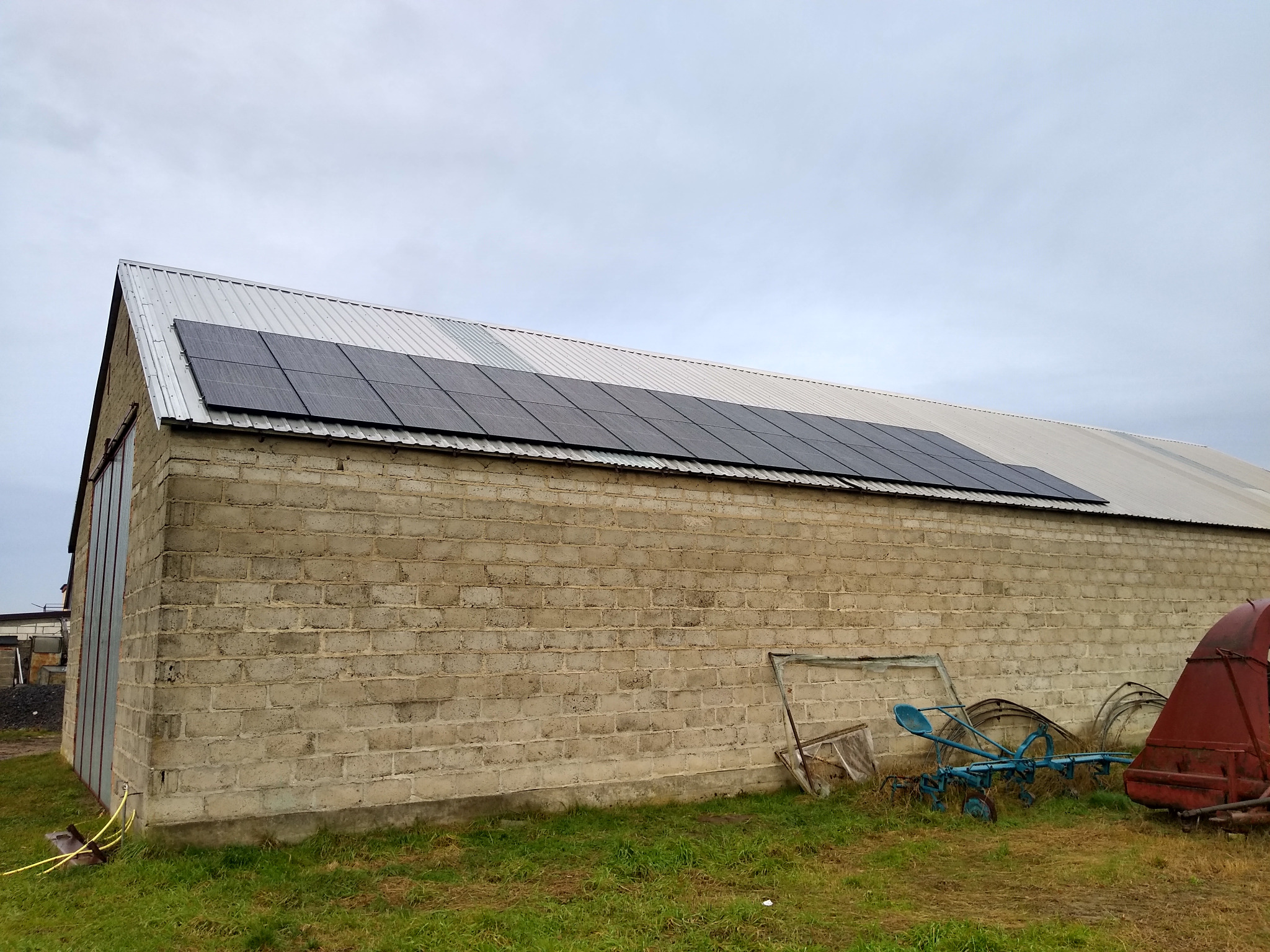 Instalacja fotowoltaiczna na dachu budynku gospodarczego z cegły, widoczne stare maszyny rolnicze w tle.