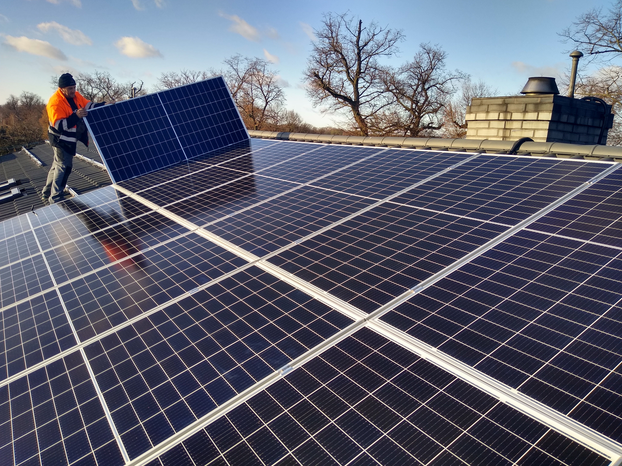 Montaż paneli słonecznych na dachu domu; pracownik w odblaskowej kurtce umieszcza panel na konstrukcji, widoczny komin i drzewa w tle.