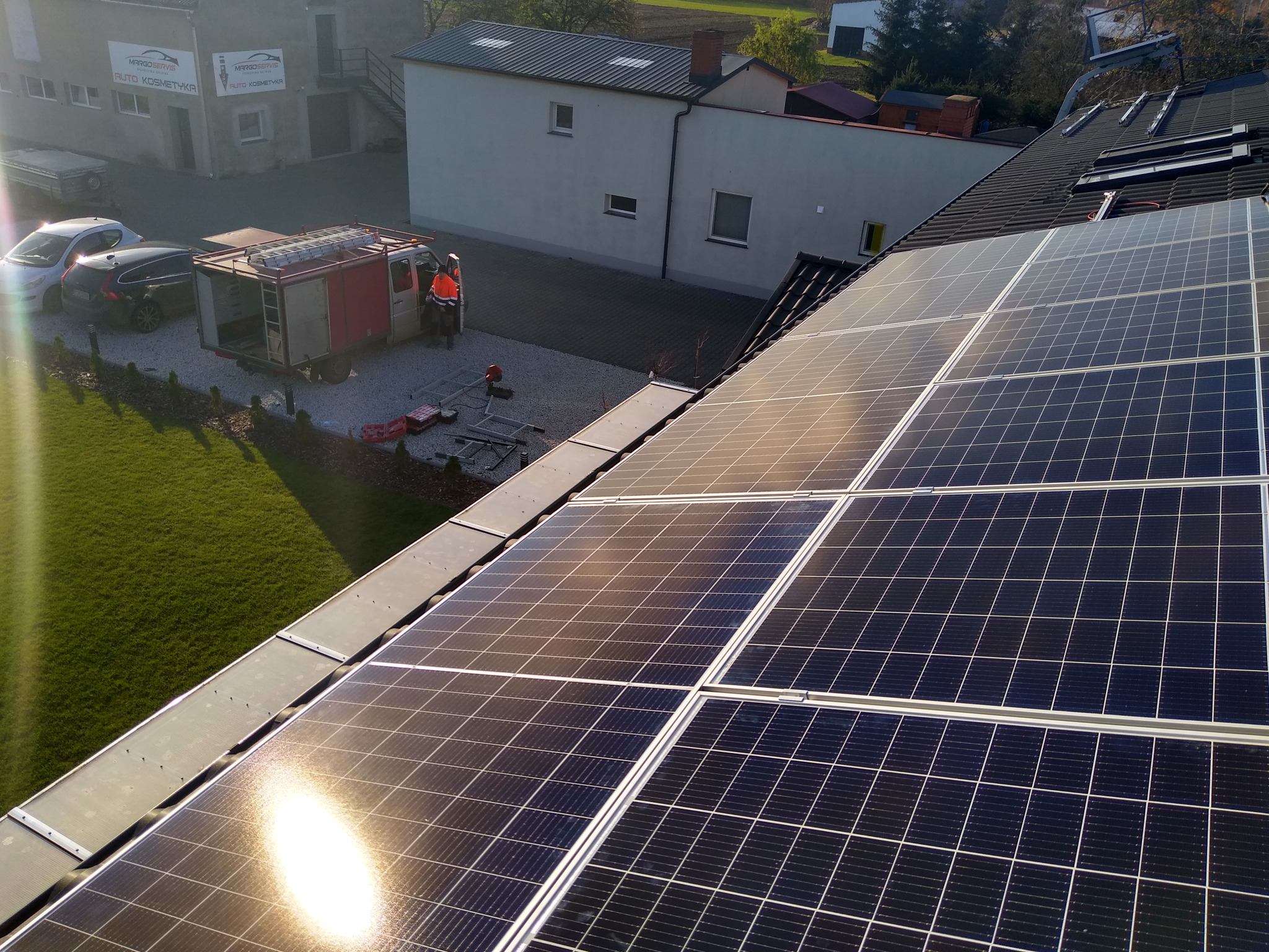Montaż paneli słonecznych na dachu domu jednorodzinnego, widoczny samochód serwisowy i pracownicy w odblaskowych kamizelkach.