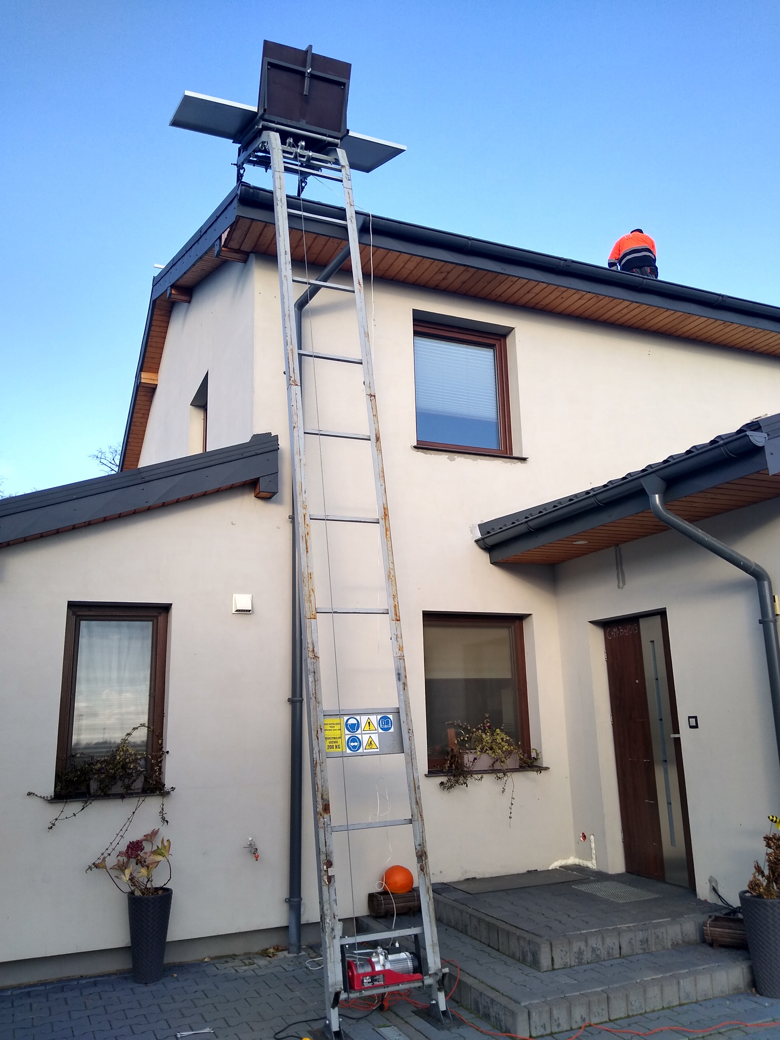 Montaż paneli słonecznych na dachu domu jednorodzinnego przy użyciu windy dekarskiej, widoczny pracownik w pomarańczowej kamizelce na dachu.