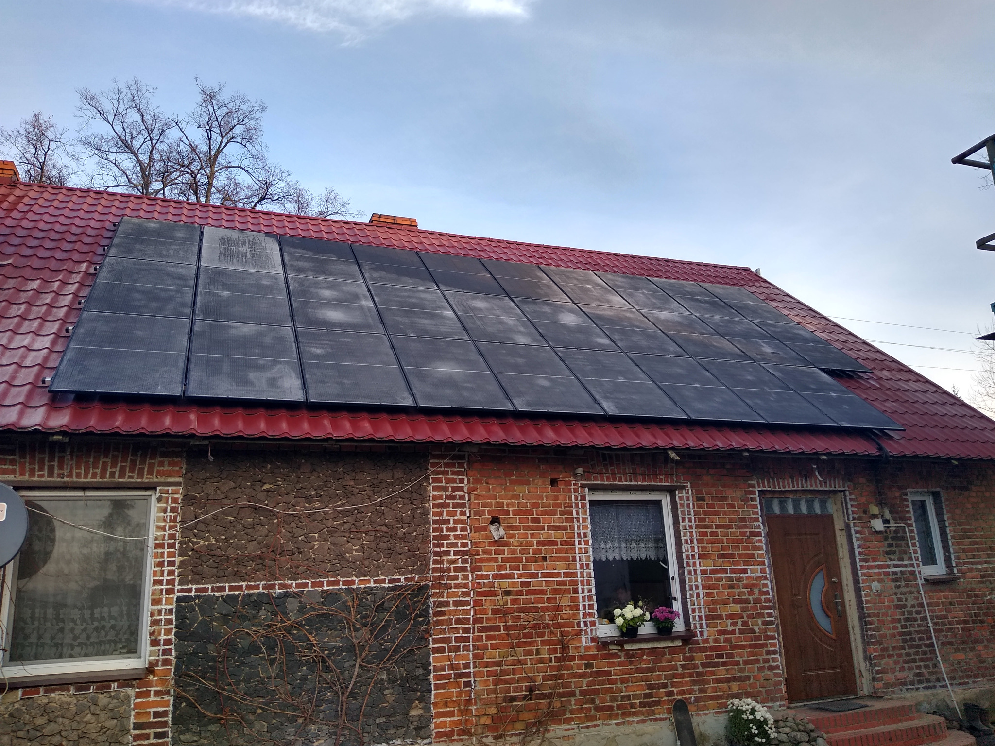 Panele słoneczne na czerwonym dachu ceglanego domu z zakrytym oknem i kwiatami w doniczkach.