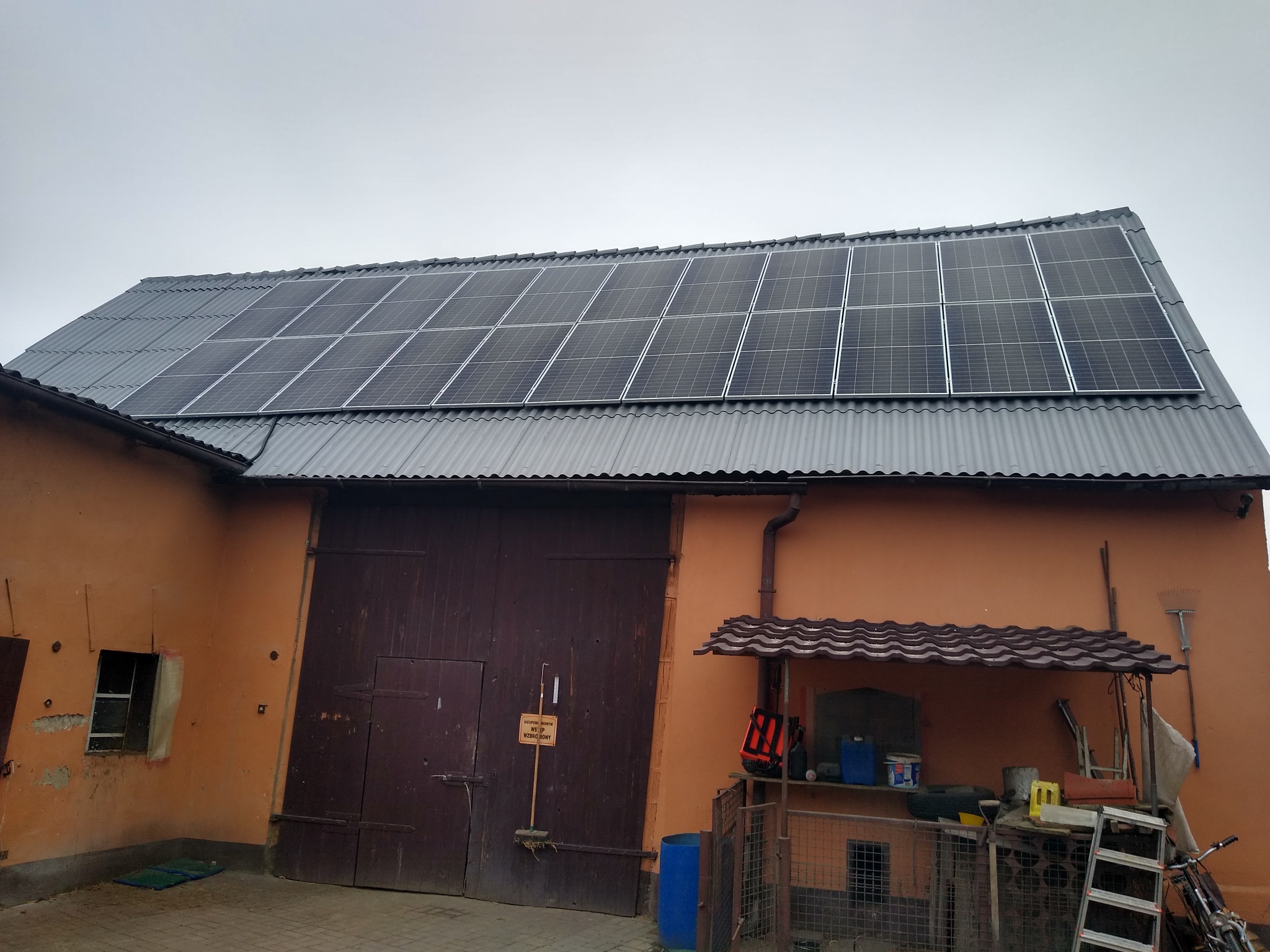 Panele słoneczne na dachu budynku gospodarczego z cegły, widoczne narzędzia i sprzęty w otoczeniu.