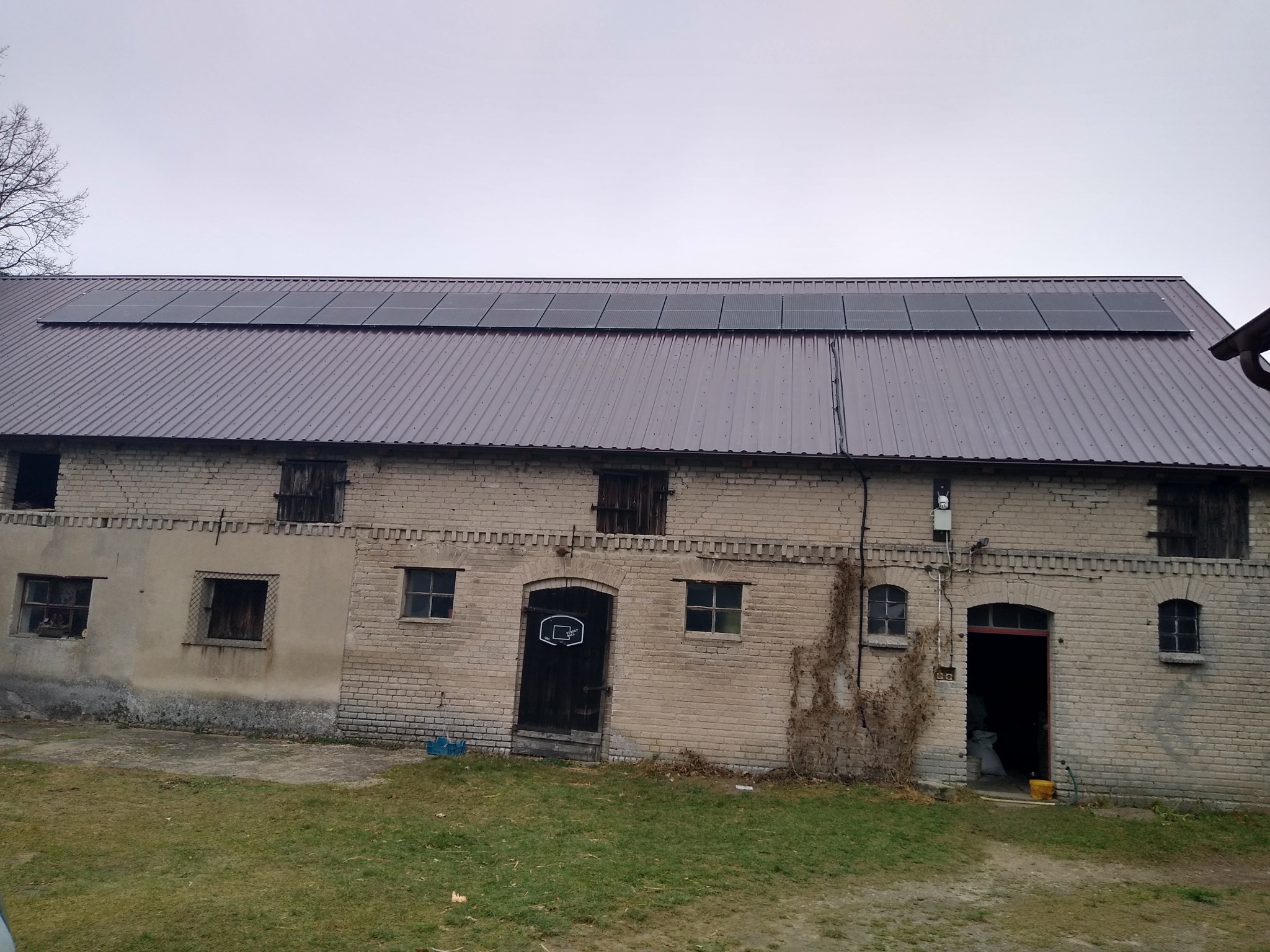 Panele słoneczne na dachu starego, ceglanego budynku gospodarczego z metalowym dachem, widoczne pnącza na ścianie i zarośnięte podwórze.