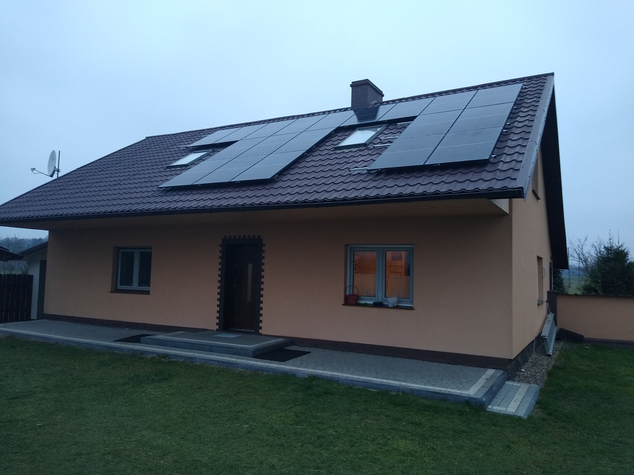 Dom jednorodzinny z brązowym dachem pokrytym panelami fotowoltaicznymi i oknami dachowymi, widok z trawnika w pochmurny dzień.