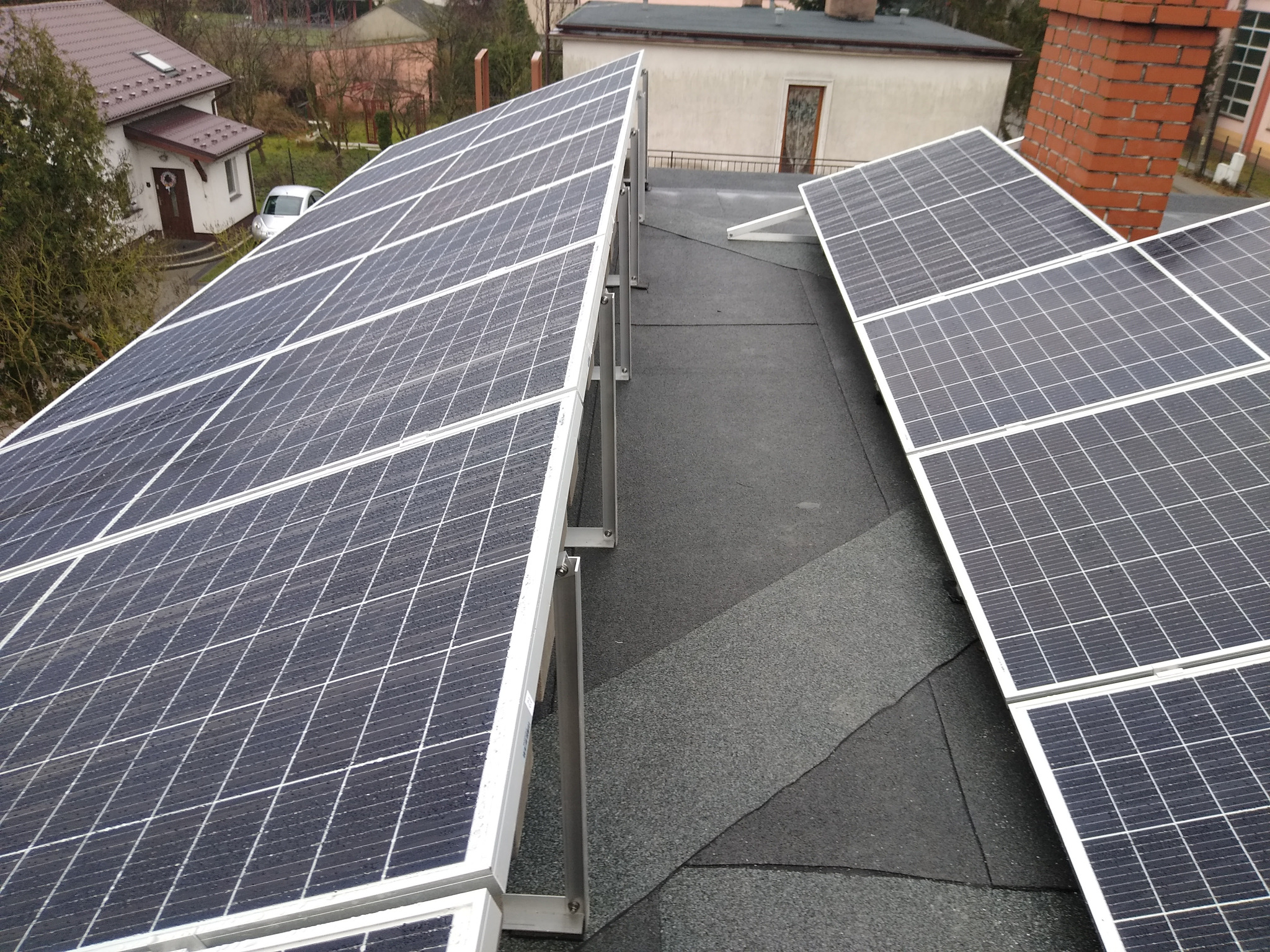 Instalacja paneli słonecznych na płaskim dachu pokrytym papą, widoczne metalowe wsporniki i krople wody na panelach.