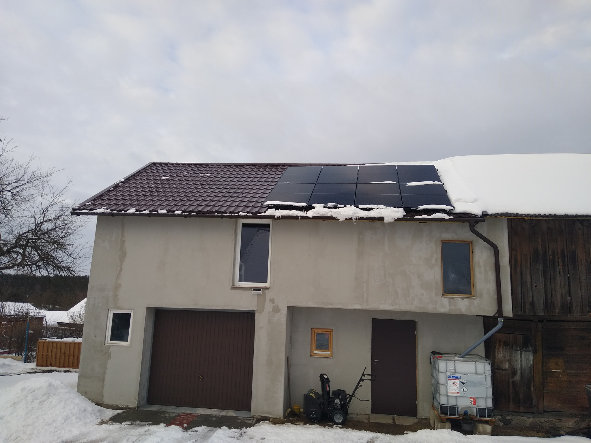 Częściowo ośnieżone panele na brązowym dachu dwukondygnacyjnego budynku z garażem i zbiornikiem na wodę deszczową. Na pierwszym planie resztki śniegu.