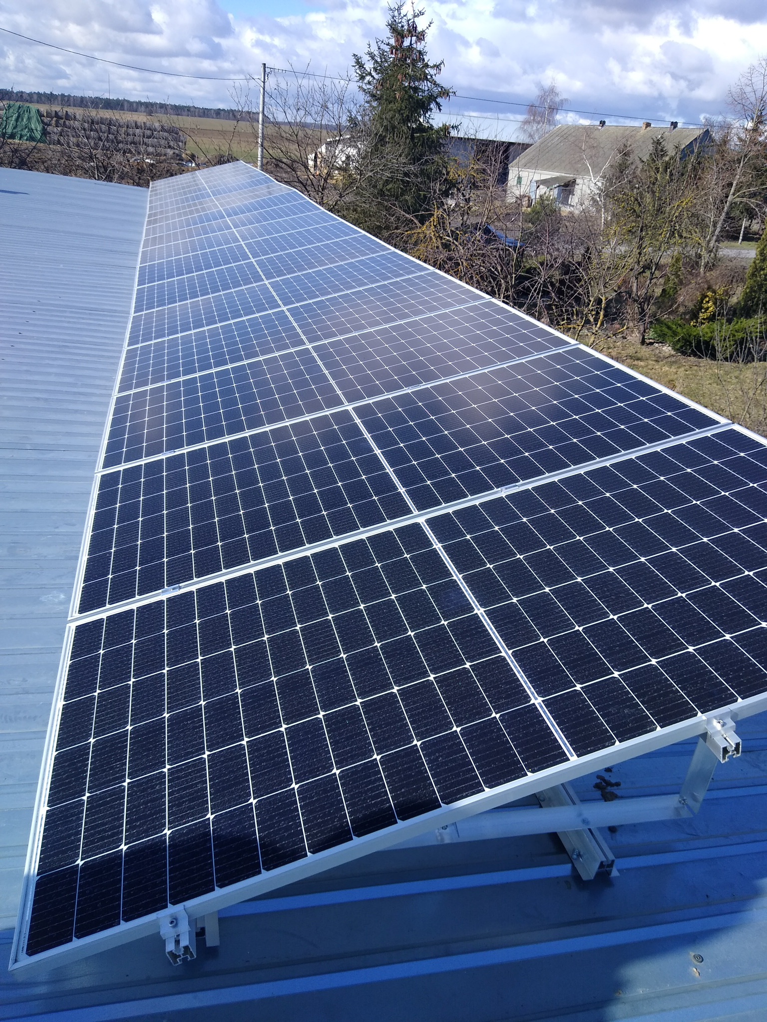 Rząd paneli słonecznych na metalowym dachu, widok z góry, z elementami montażowymi i tłem z drzewami i budynkami w oddali.