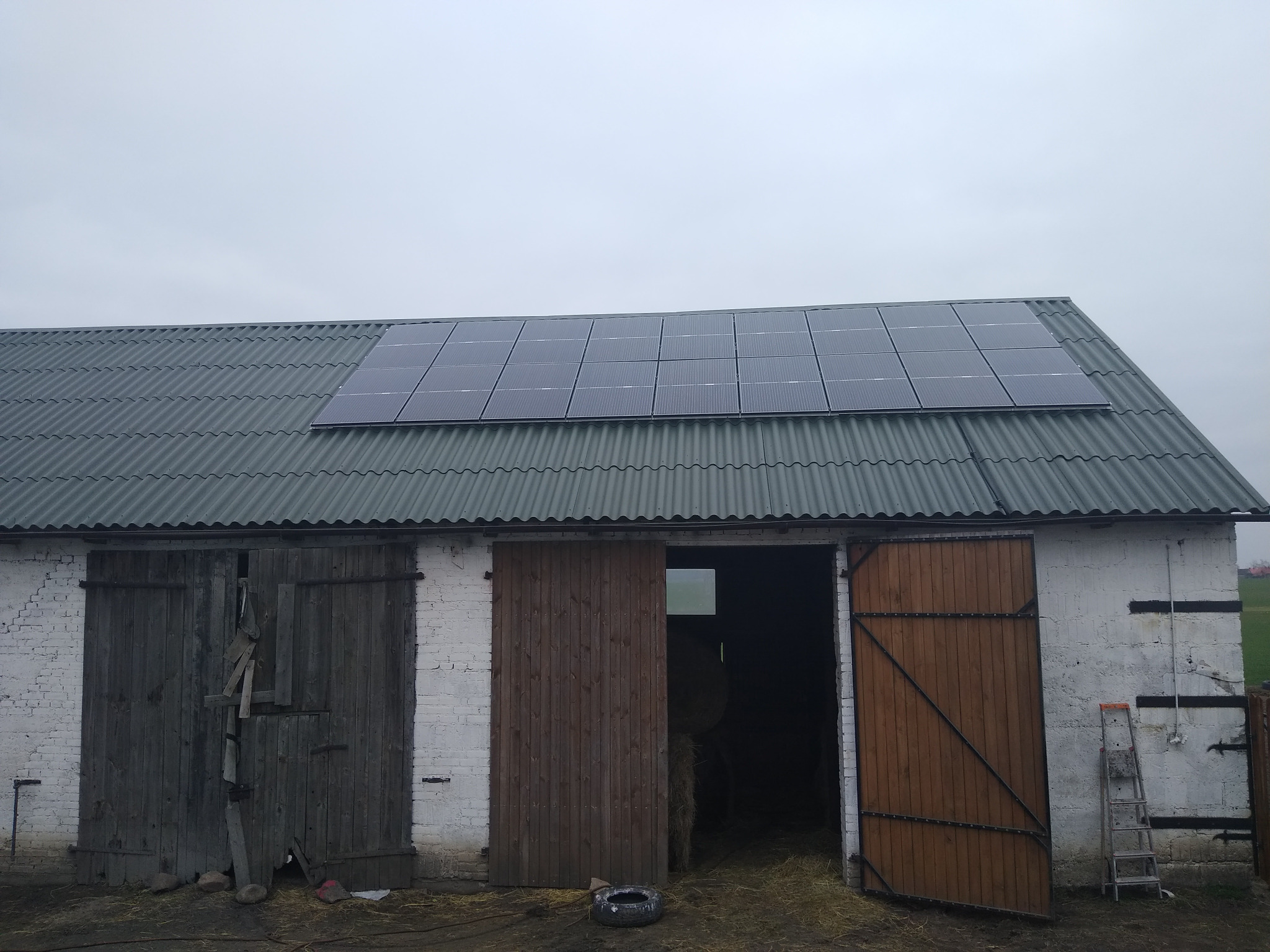 Instalacja paneli na dachu budynku gospodarczego z otwartymi drewnianymi wrotami i drabiną opartą o ścianę.