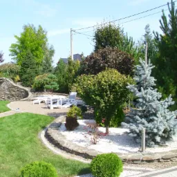 Urozmaicony ogród z licznymi drzewami i krzewami ozdobnymi, białymi kamykami, trawnikiem, ścieżką i kamienną kaskadą w tle, z białymi leżakami ogrodowymi na górze.