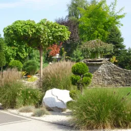 Ogród z różnorodną roślinnością: kuliste drzewa, ozdobne trawy, kamień wapienny i kamienny murek z wodospadem w tle.