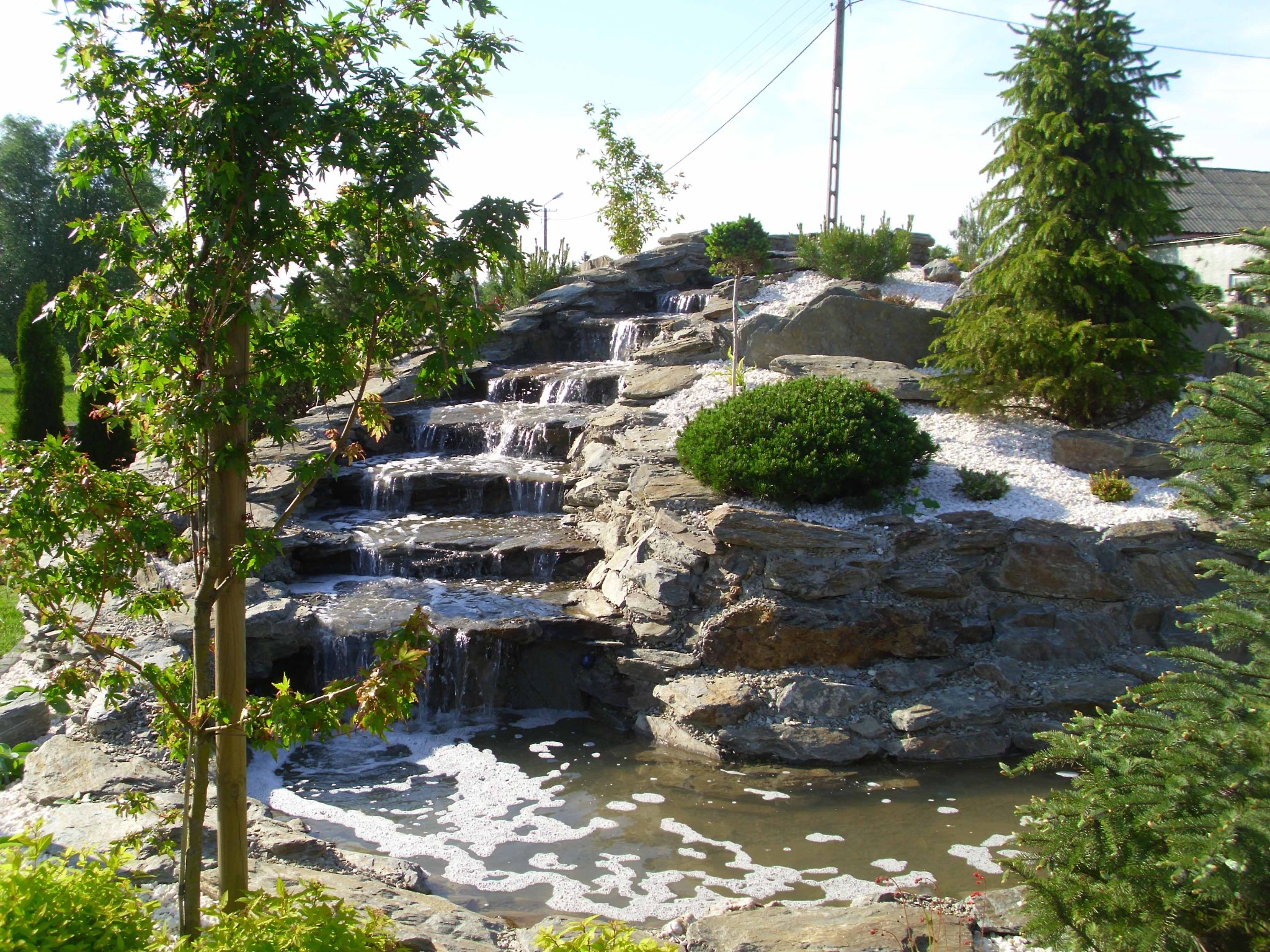 Kaskada wodna z kamieni w ogrodzie, z białym grysem, zielonymi krzewami i młodymi drzewami, w tle widoczny budynek.