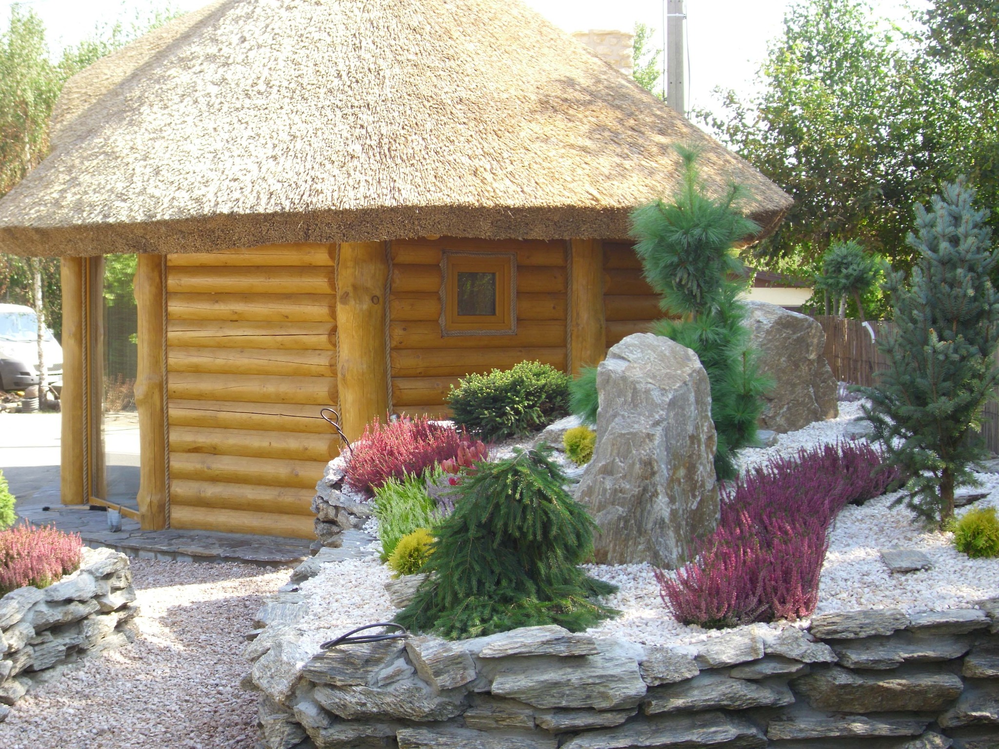 Ogród w Rypinie z drewnianą altaną krytą strzechą, ozdobiony kamieniami, wrzosami i iglakami na białym żwirze.