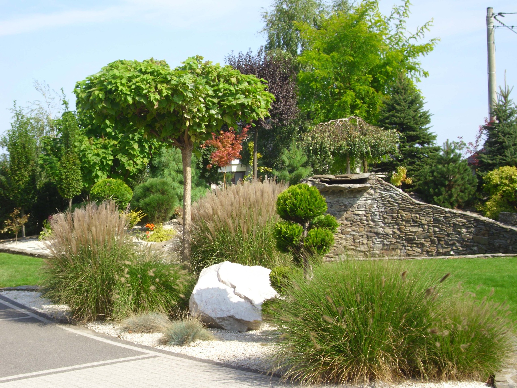 Ogród z różnorodną roślinnością: kuliste drzewa, ozdobne trawy, kamień wapienny i kamienny murek z wodospadem w tle.