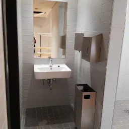 Nowoczesna łazienka publiczna z kwadratową umywalką, chromowaną baterią z sensorem, dozownikiem mydła, podajnikiem ręczników papierowych i metalowym koszem na śmieci; szare płytki na ścianach...