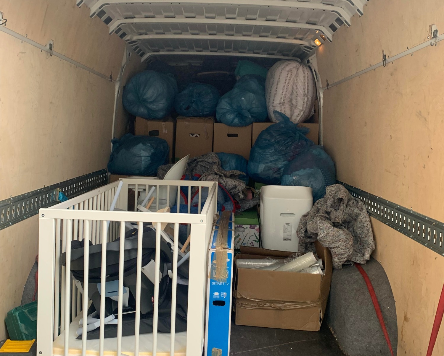 Wnętrze furgonetki wypełnionej kartonami, workami z rzeczami i rozłożonym białym łóżeczkiem dziecięcym, przygotowanymi do transportu.