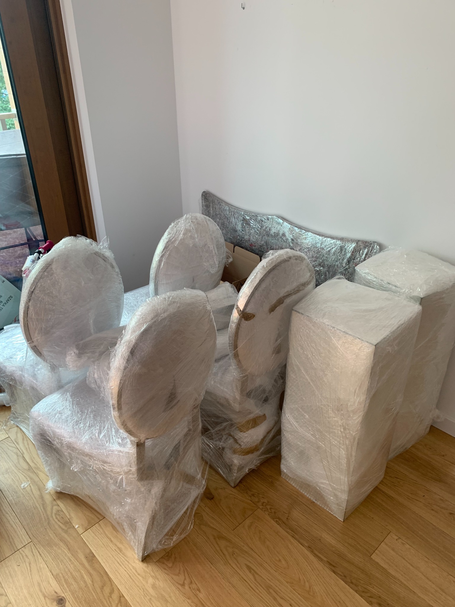Meble (krzesła z okrągłymi oparciami i szafki) owinięte folią stretch, przygotowane do transportu, ustawione w rogu pokoju na drewnianej podłodze.