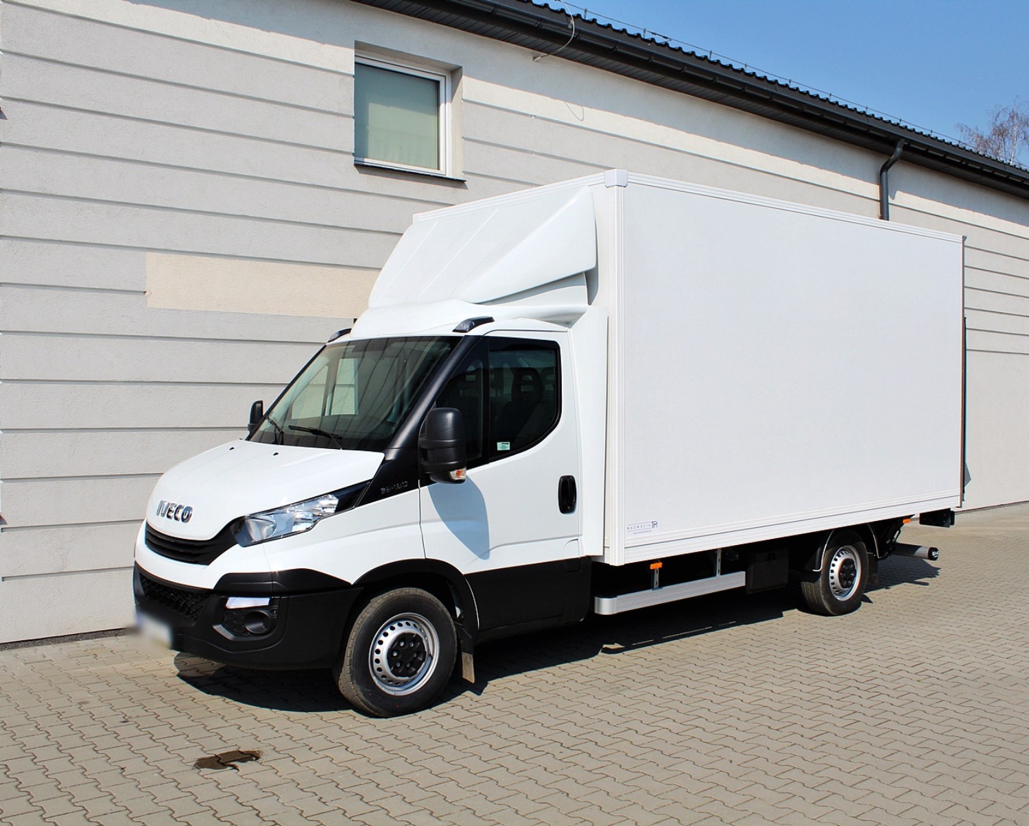 Biały samochód dostawczy marki Iveco z zabudową kontenerową na kostce brukowej przed budynkiem o jasnej elewacji w słoneczny dzień.