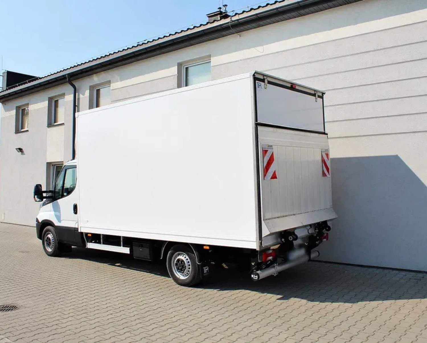 Biały samochód dostawczy z otwartą przestrzenią ładunkową zaparkowany przed budynkiem z szarą elewacją i kostką brukową.