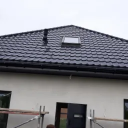 Nowy, czarny dach z dachówki modułowej z oknem dachowym i kominkiem wentylacyjnym, widok z dołu na tle szarego nieba, widoczne rusztowania przy ścianie budynku.