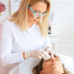 Katarzyna von Engel - makijaż permanentny brwi (microblading) Warszawa - Kosmetyczka w okularach, w białym uniformie i rękawiczkach, oczyszcza brwi klientki leżącej na białym prześcieradle, w tle stolik z hortensjami w wazonie.