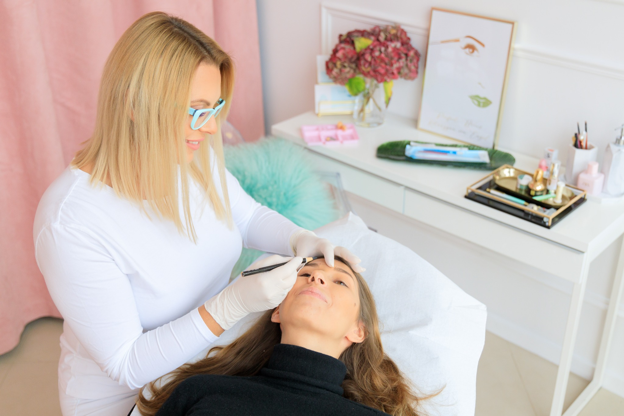 Kobieta w białej bluzce i turkusowych okularach, w lateksowych rękawiczkach, rysuje brwi klientce leżącej na białym łóżku kosmetycznym, w tle biała toaletka z kwiatami i akcesoriami.