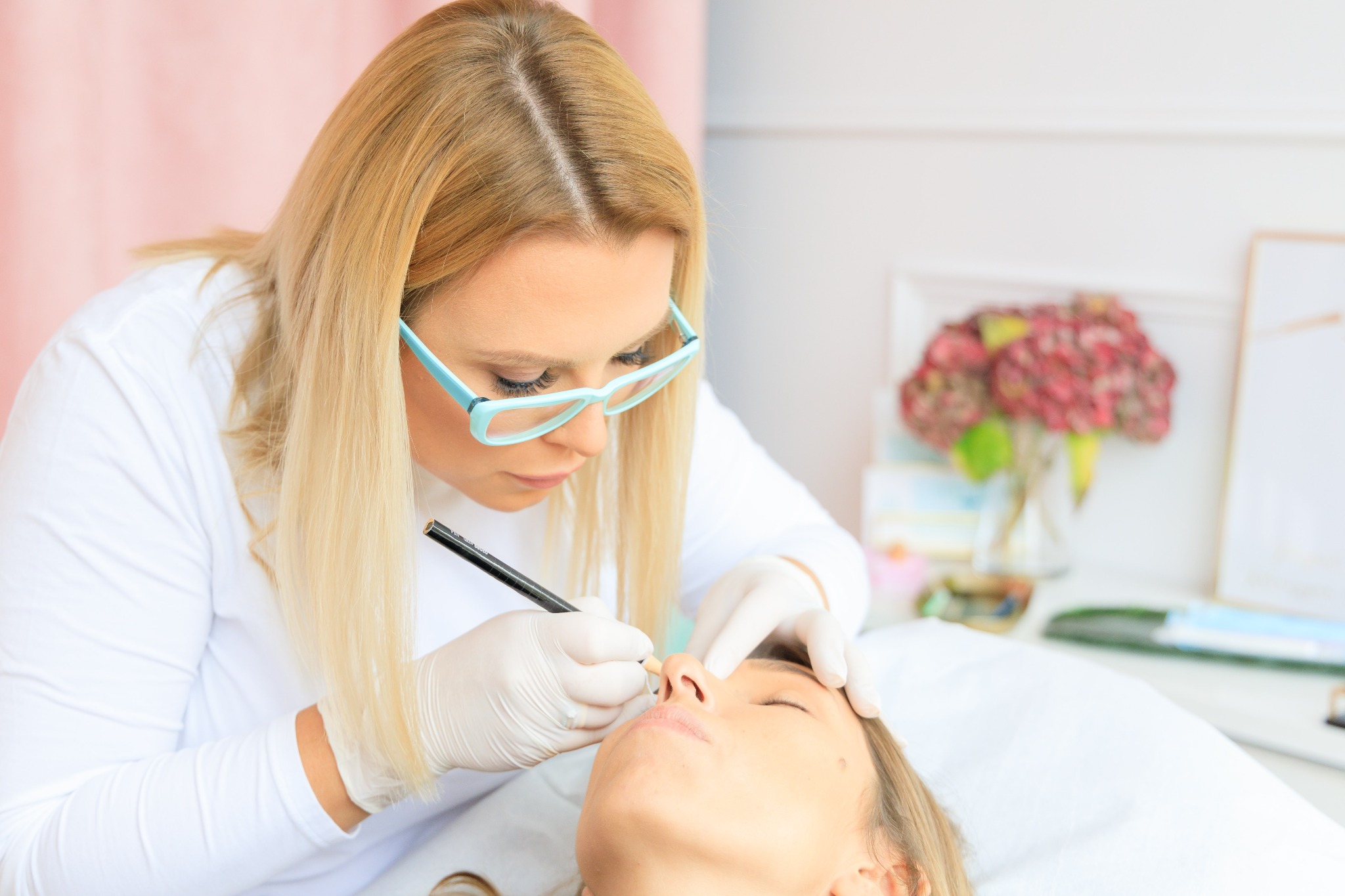 Blond kosmetolog w niebieskich okularach i białych rękawiczkach wykonuje precyzyjny makijaż permanentny brwi klientce leżącej na białym łóżku kosmetycznym, używając czarnego pisaka.