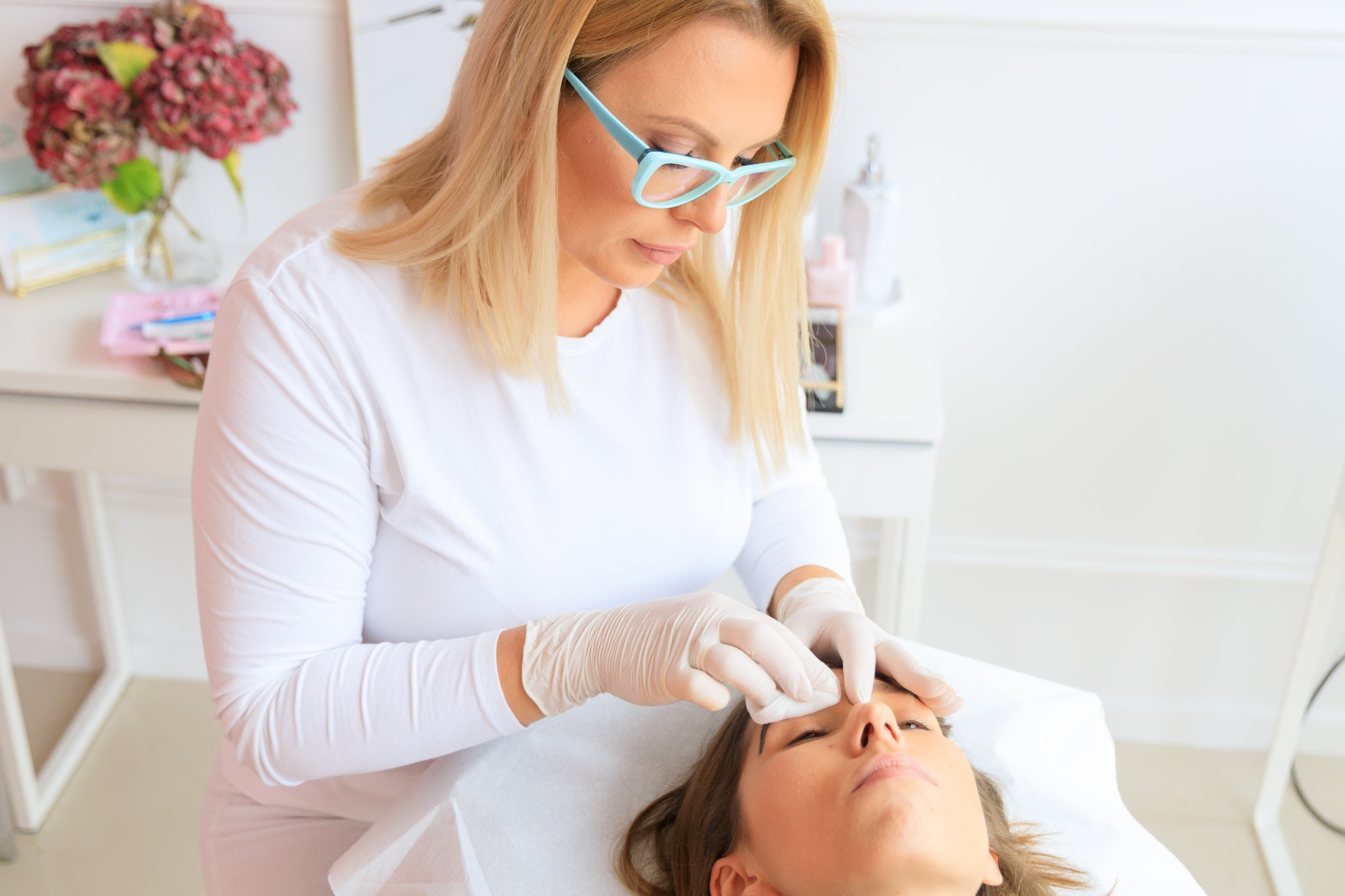 Kosmetyczka w okularach, w białym uniformie i rękawiczkach, oczyszcza brwi klientki leżącej na białym prześcieradle, w tle stolik z hortensjami w wazonie.
