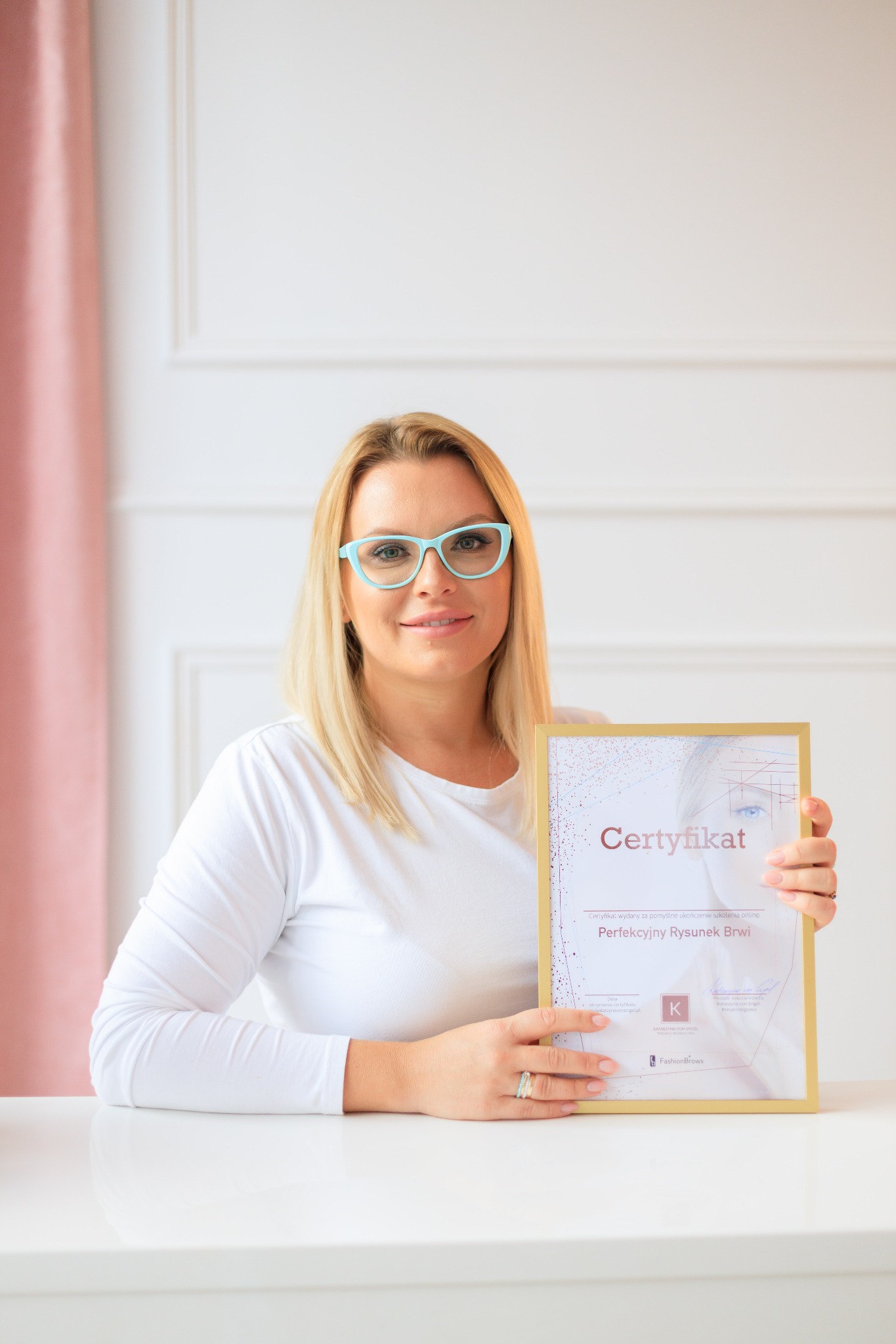Blondynka w turkusowych okularach prezentuje certyfikat ukończenia szkolenia online z perfekcyjnego rysunku brwi, trzymając go w dłoniach na tle białego stołu i ściany z dekoracyjną sztukaterią.
