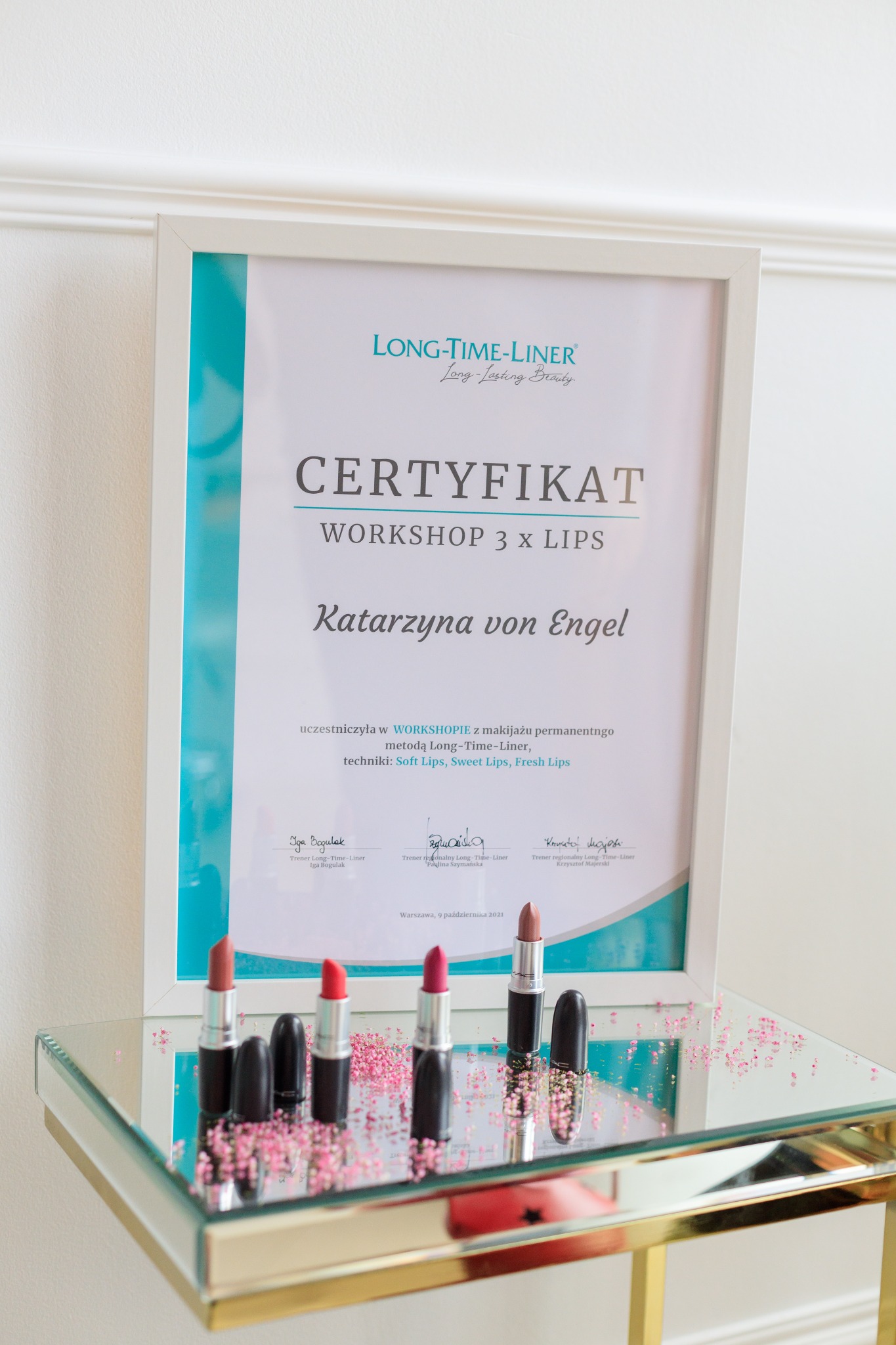 Certyfikat ukończenia warsztatów makijażu permanentnego ust Long-Time-Liner, otwarte i zamknięte szminki w odcieniach czerwieni i różu na szklanym stoliku z dekoracją z drobnych różowych kwiatków.