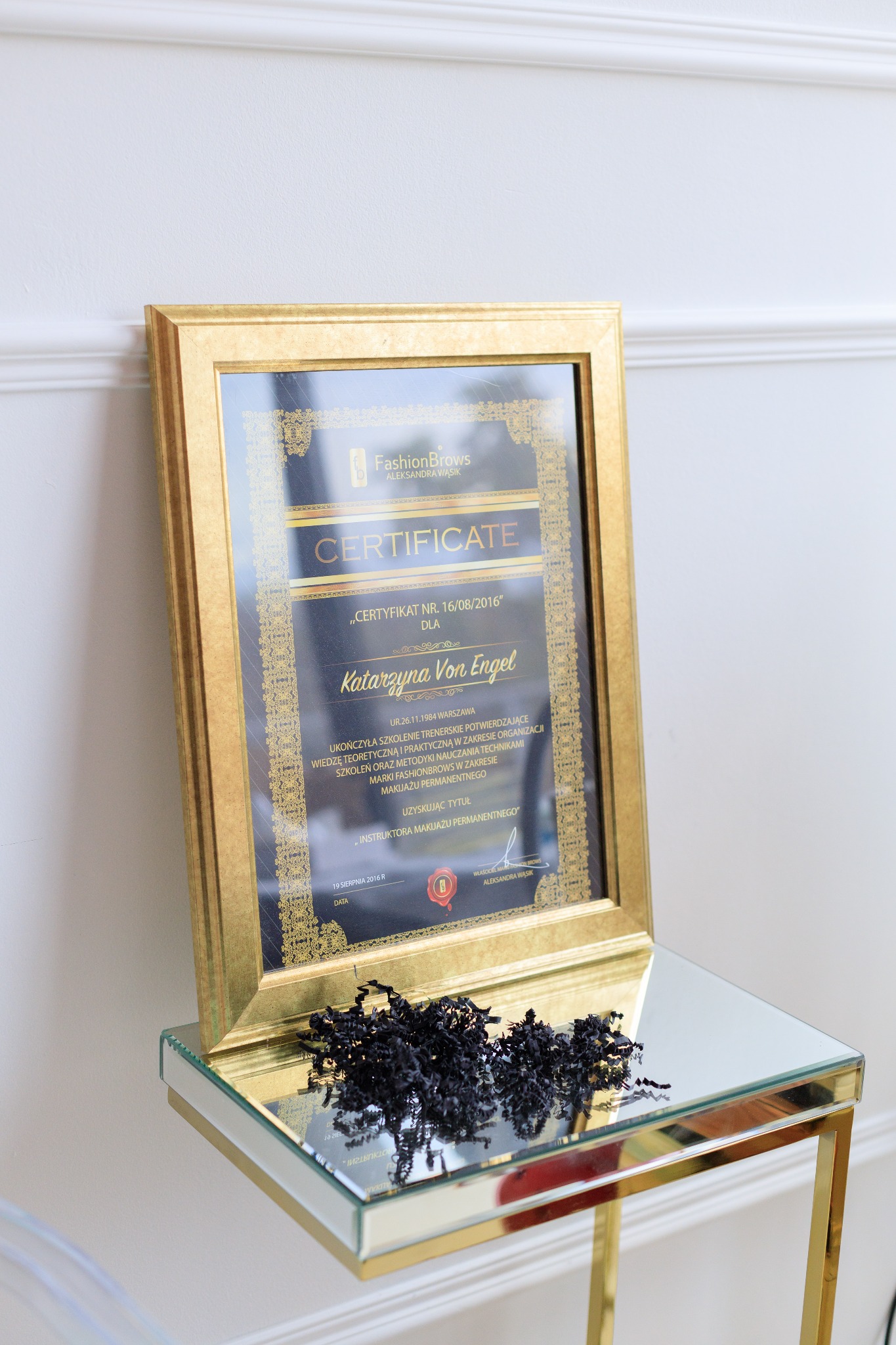 Złota ramka z certyfikatem FashionBrows dla Katarzyny Von Engel, potwierdzającym ukończenie szkolenia trenerskiego w zakresie makijażu permanentnego, leżąca na lustrzanym stoliku obok czarnego...