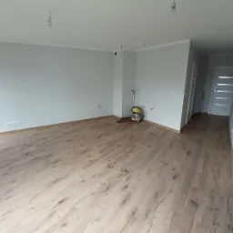 Wnętrze pokoju po remoncie z drewnianą podłogą, szarymi ścianami i widoczną szlifierką do parkietu w rogu pomieszczenia.