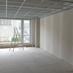 Wnętrze w trakcie prac wykończeniowych: ściany z nałożoną gładzią, metalowa konstrukcja sufitu podwieszanego i drabina aluminiowa przy oknie.