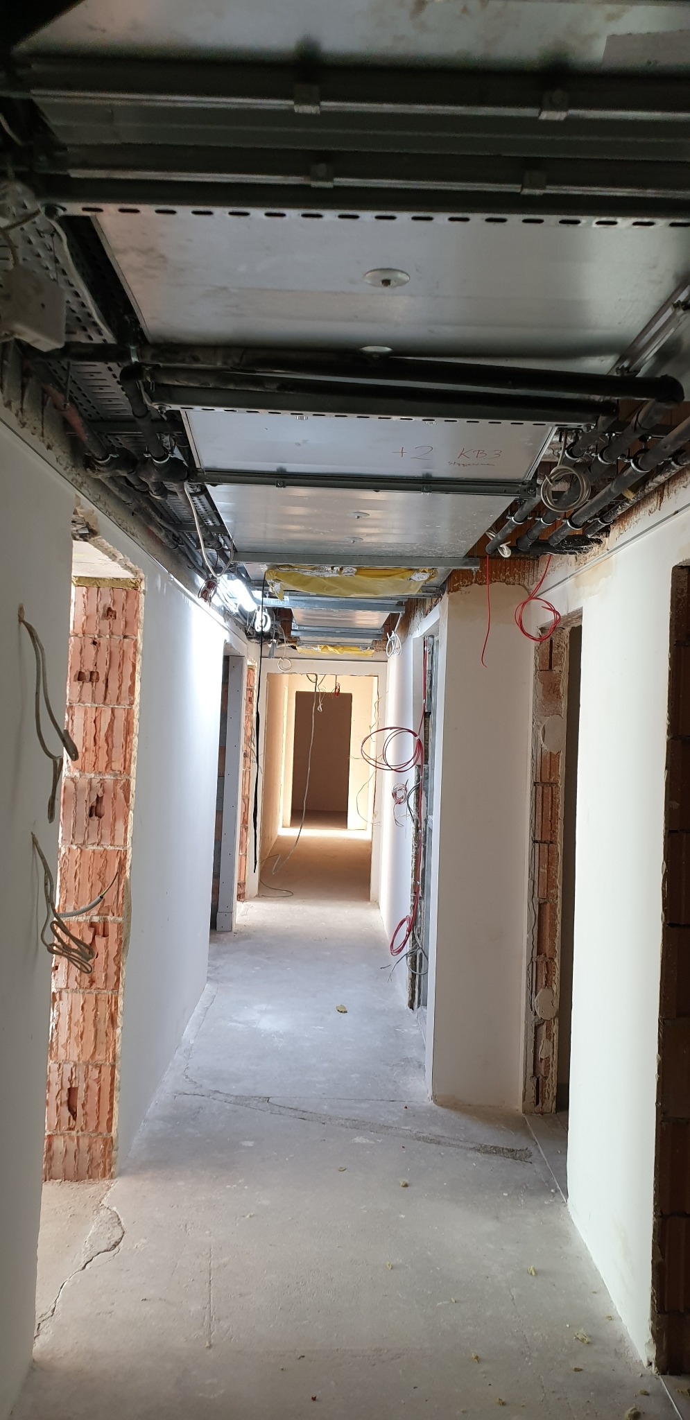 Korytarz w trakcie remontu z widoczną surową cegłą, instalacjami pod sufitem i przygotowaną podłogą.
