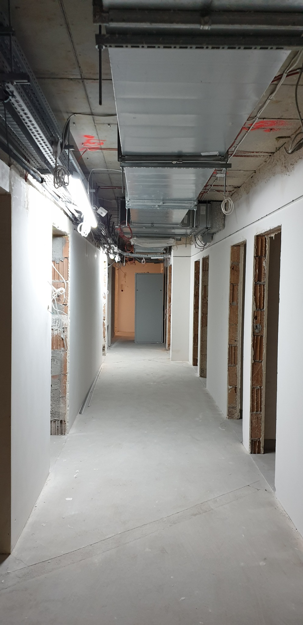 Korytarz w trakcie prac wykończeniowych, widoczne surowe ściany z fragmentami cegieł, świeżo położona gładź, metalowe kanały wentylacyjne pod sufitem oraz instalacje elektryczne.
