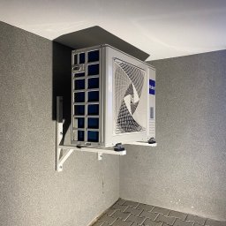 Mantex Instalacje - Zamontowana jednostka zewnętrzna klimatyzacji Haier na wspornikach, widoczna z dołu w rogu pomieszczenia z szarą ścianą i podłogą z kostki brukowej.