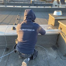 Mantex Instalacje - Pracownik w odzieży roboczej z napisem 'Klimatyzacja' instaluje system na dachu we Wrocławiu. Widoczne przewody i elementy montażowe.