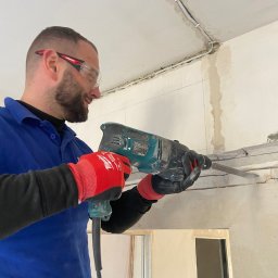 Mantex Instalacje - Pracownik w okularach wierci bruzdy w ścianie pod kable. Widoczne czerwone rękawice i niebieska bluza robocza. W tle instalacja elektryczna pod sufitem.