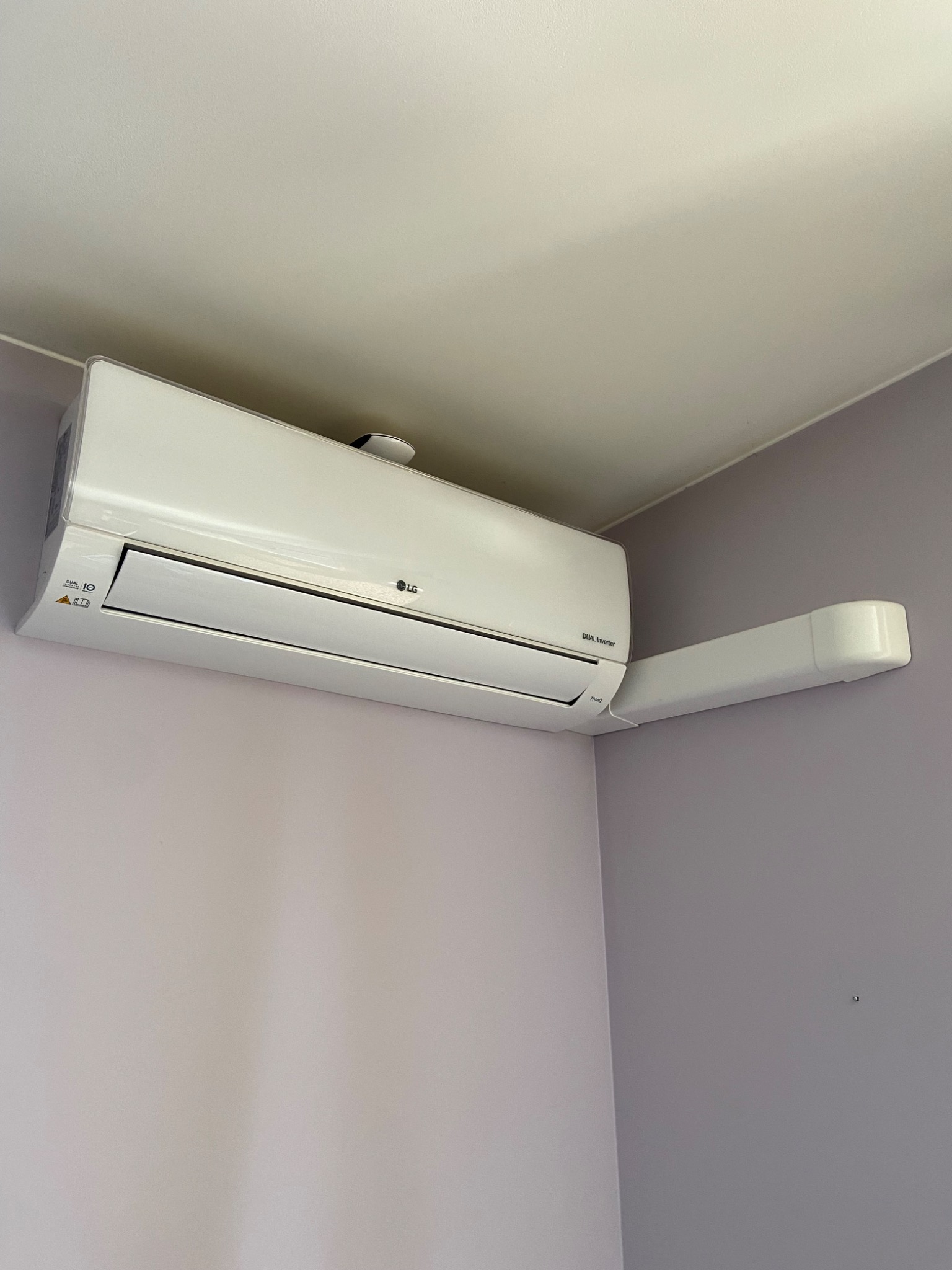 Biały klimatyzator LG Dual Inverter zamontowany na ścianie pomalowanej na dwa odcienie fioletu, z widocznym przewodem odprowadzającym skropliny.