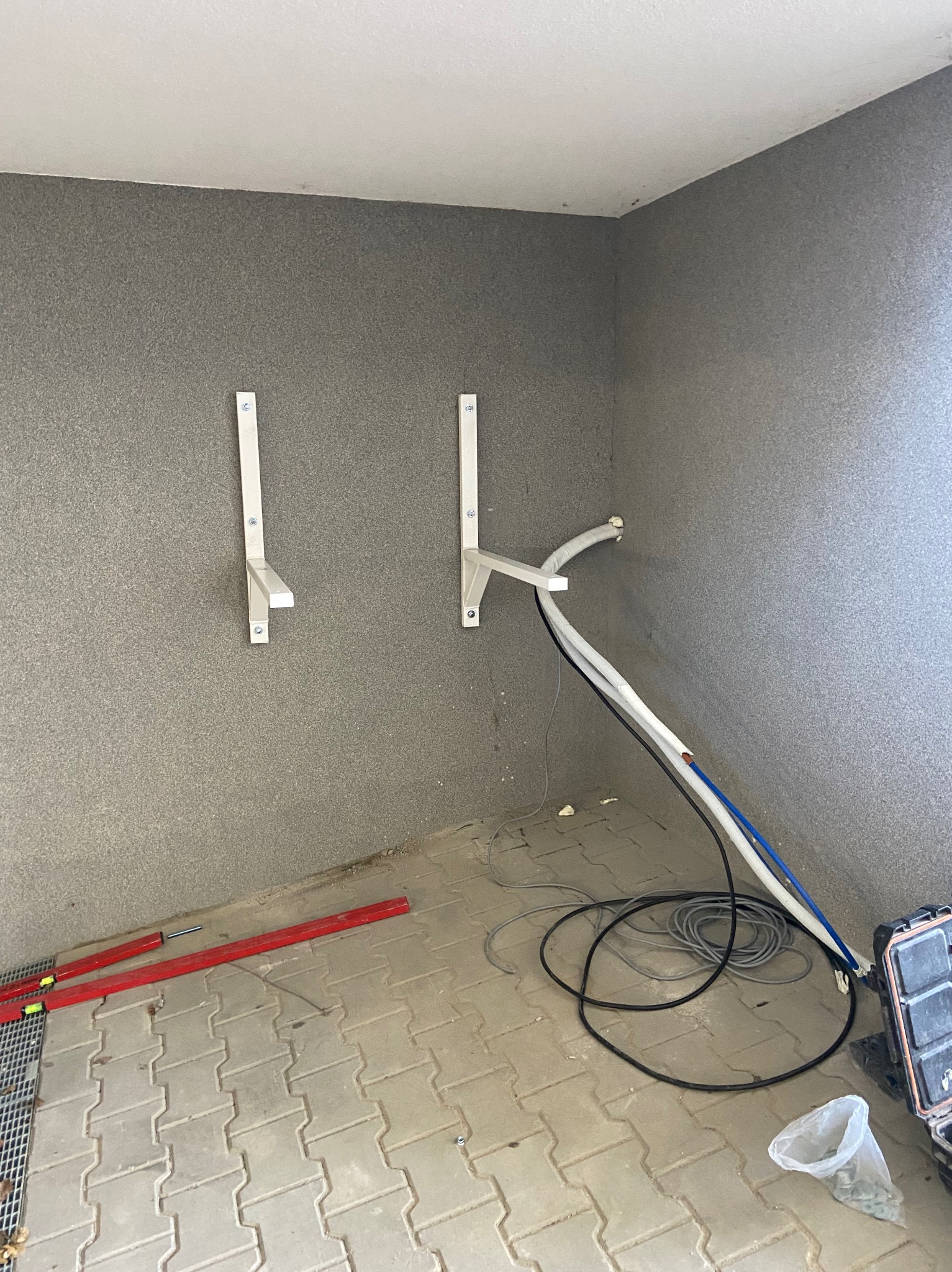 Przygotowanie do montażu klimatyzacji: wsporniki, przewody i narzędzia na podłodze w szarym pomieszczeniu. Etap instalacji przed montażem jednostki zewnętrznej.