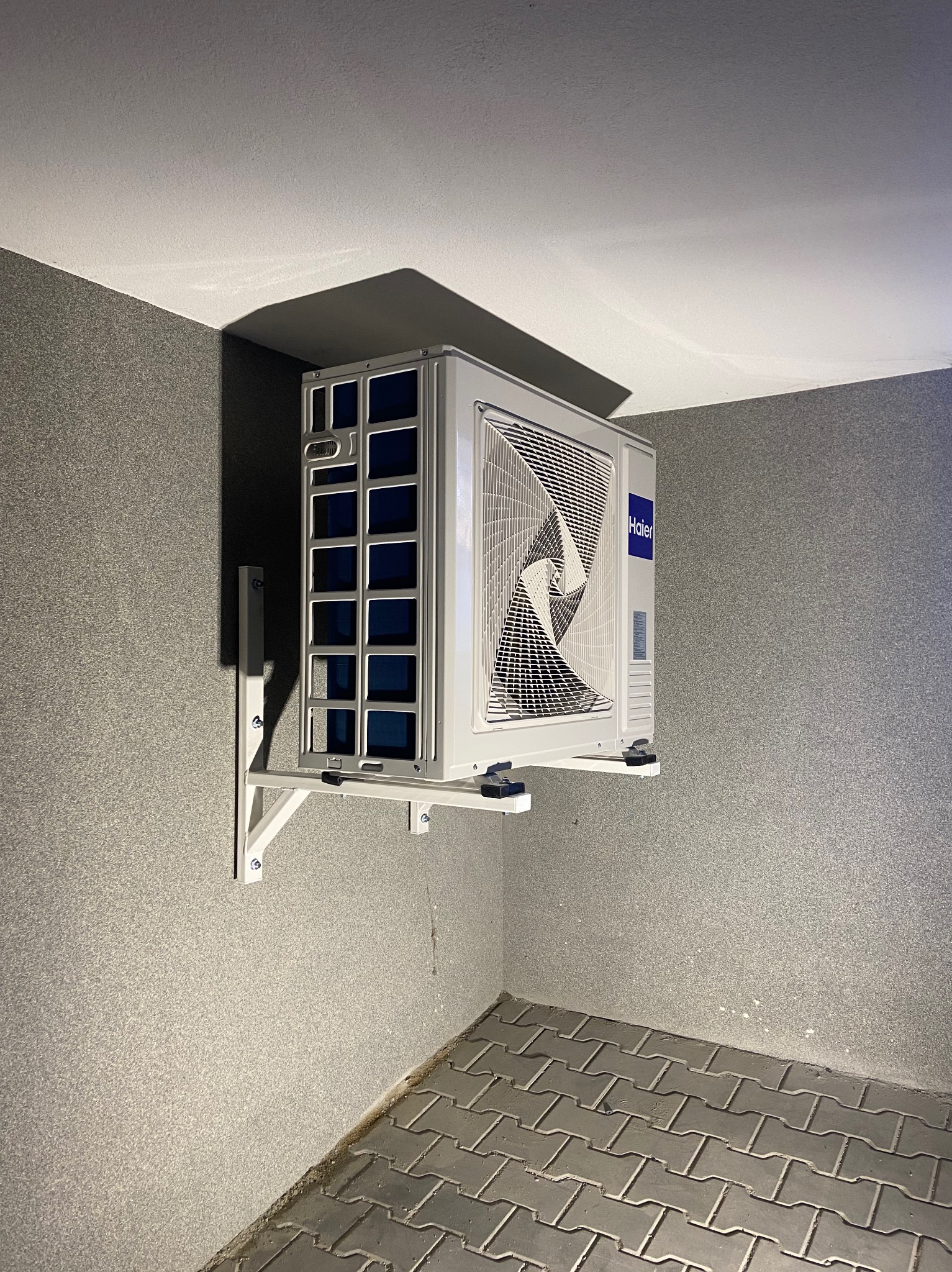 Zamontowana jednostka zewnętrzna klimatyzacji Haier na wspornikach, widoczna z dołu w rogu pomieszczenia z szarą ścianą i podłogą z kostki brukowej.