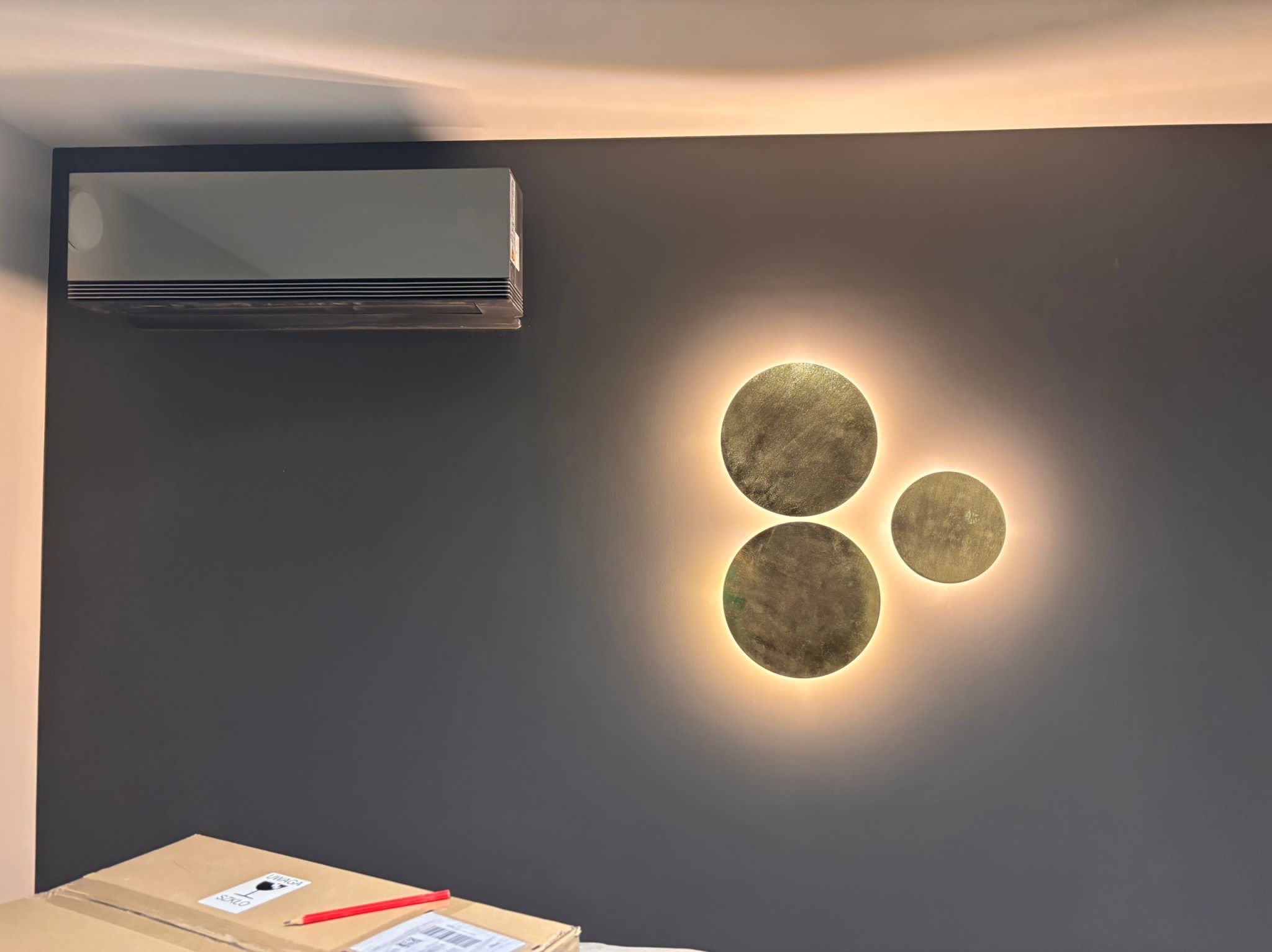 Nowoczesny klimatyzator ścienny LG na szarej ścianie z dekoracyjnym oświetleniem. Częściowo widoczny karton z akcesoriami montażowymi na dole kadru.