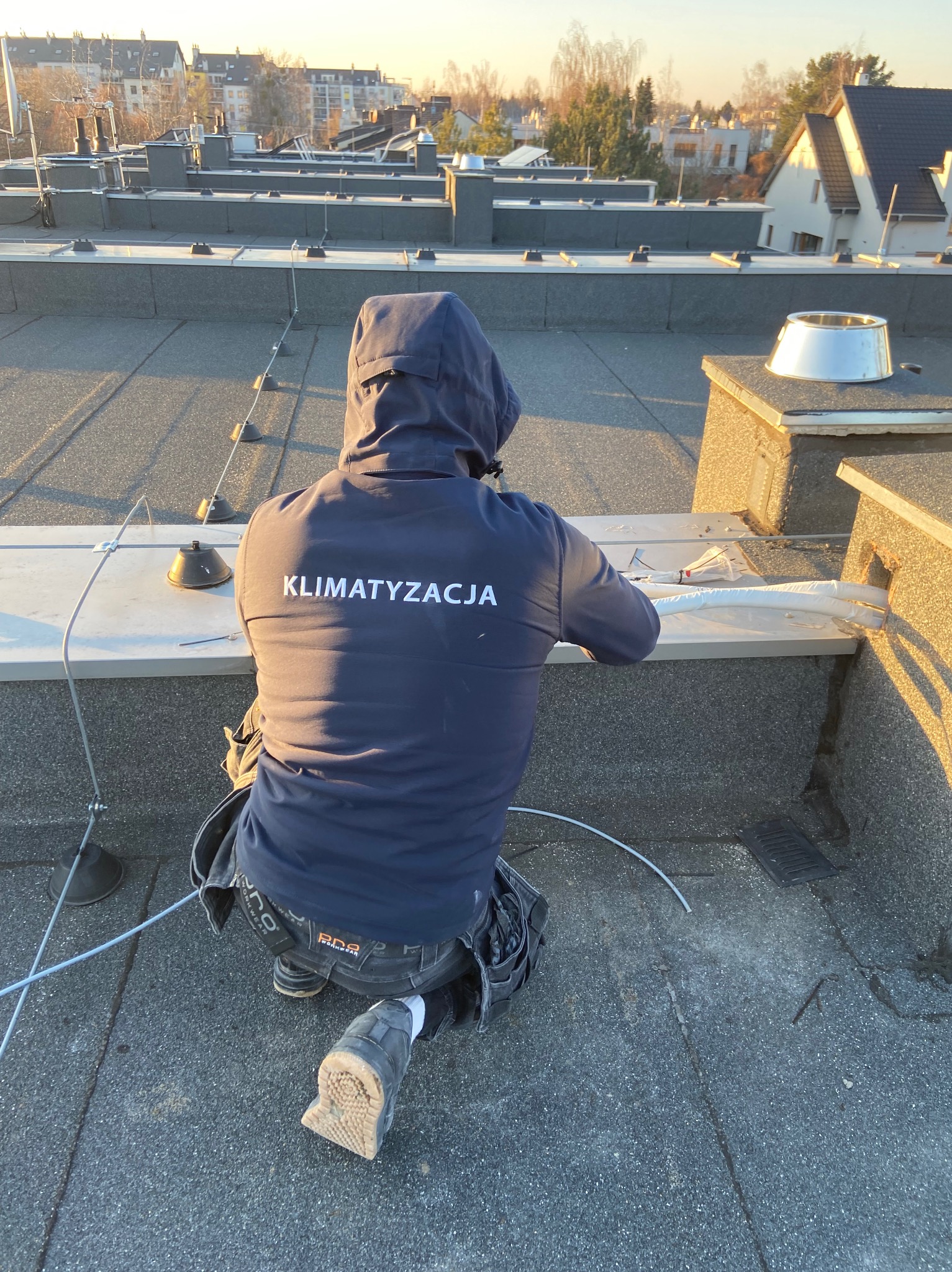 Pracownik w odzieży roboczej z napisem 'Klimatyzacja' instaluje system na dachu we Wrocławiu. Widoczne przewody i elementy montażowe.