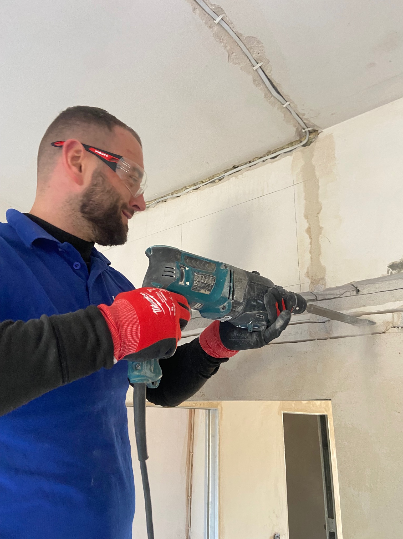 Pracownik w okularach wierci bruzdy w ścianie pod kable. Widoczne czerwone rękawice i niebieska bluza robocza. W tle instalacja elektryczna pod sufitem.